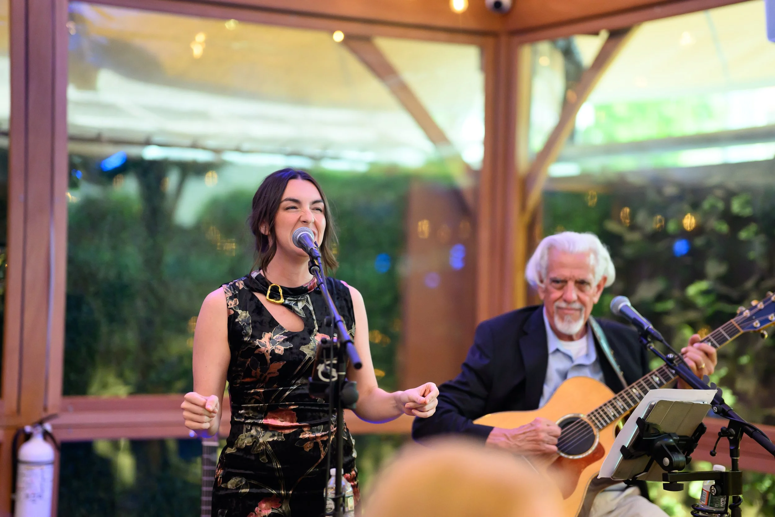 A young woman singing into a microphone at an outdoor performance, with an older man playing guitar beside her. They are under a wooden structure with greenery in the background.
