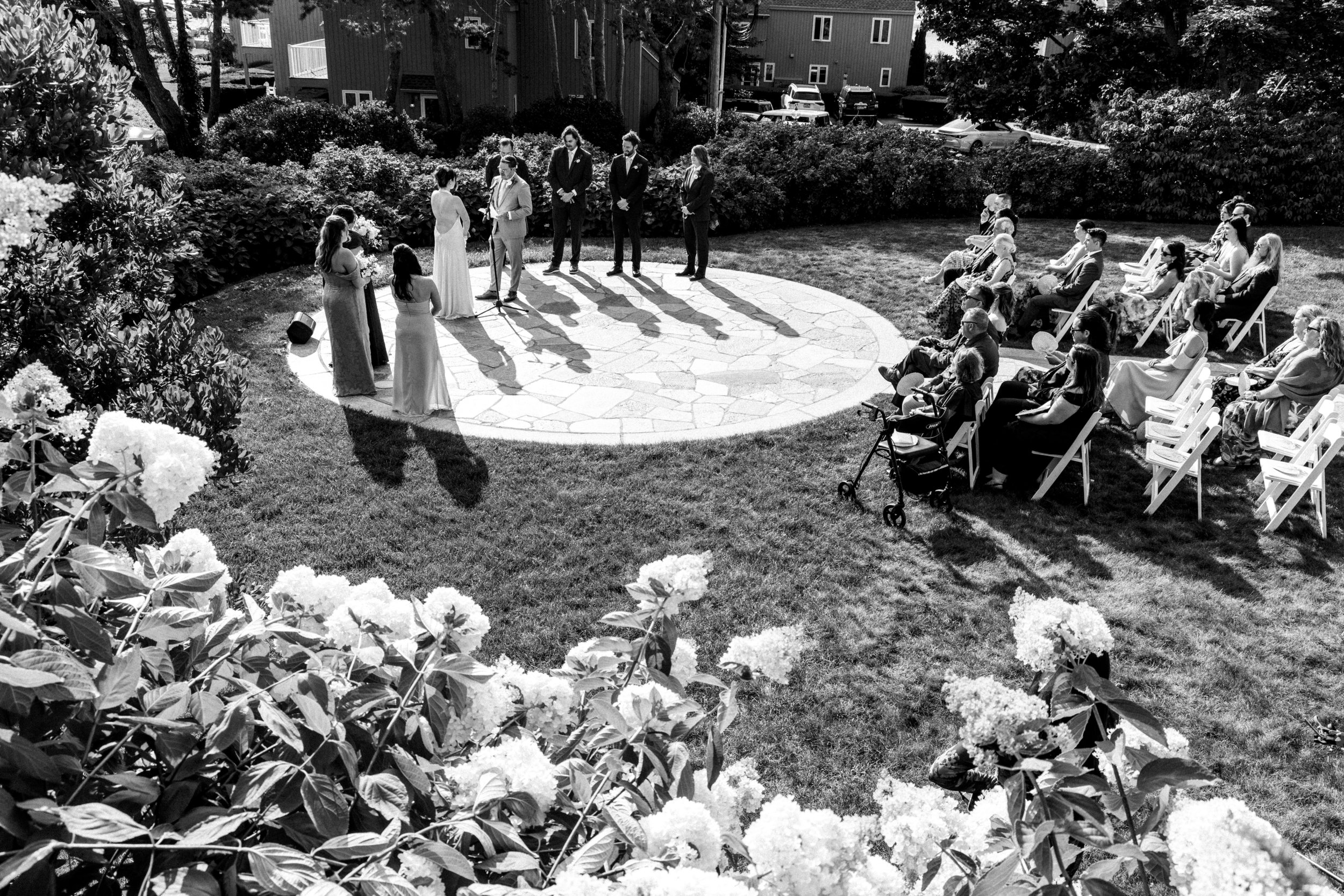 A black and white photo of an outdoor wedding ceremony in a garden with guests seated on chairs, and a couple standing and facing each other in front of an officiant. The ceremony takes place on a circular paved area surrounded by flowers and bushes.