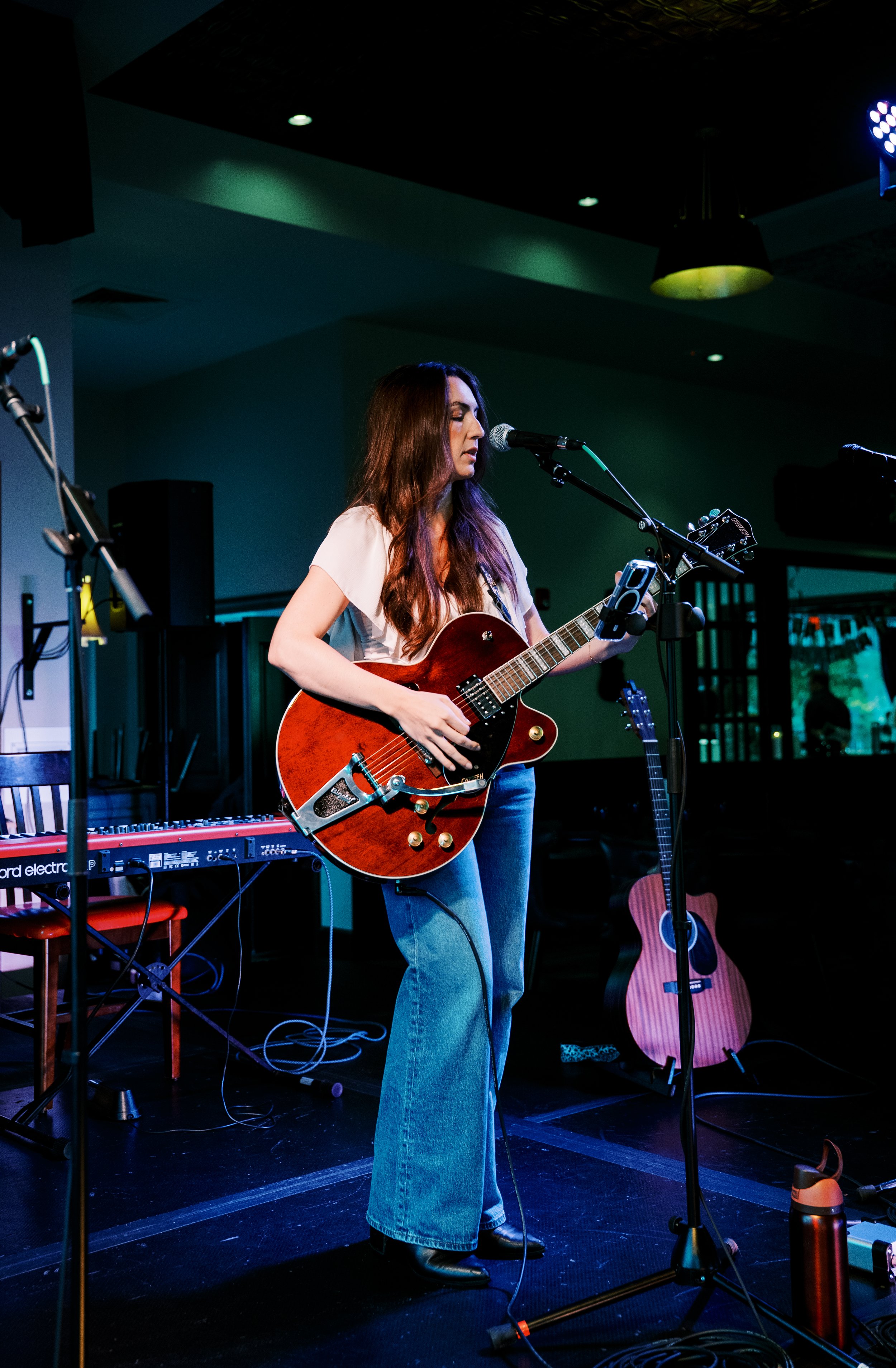 A woman with long, wavy hair is playing an acoustic guitar and singing into a microphone on a stage with musical equipment in the background.