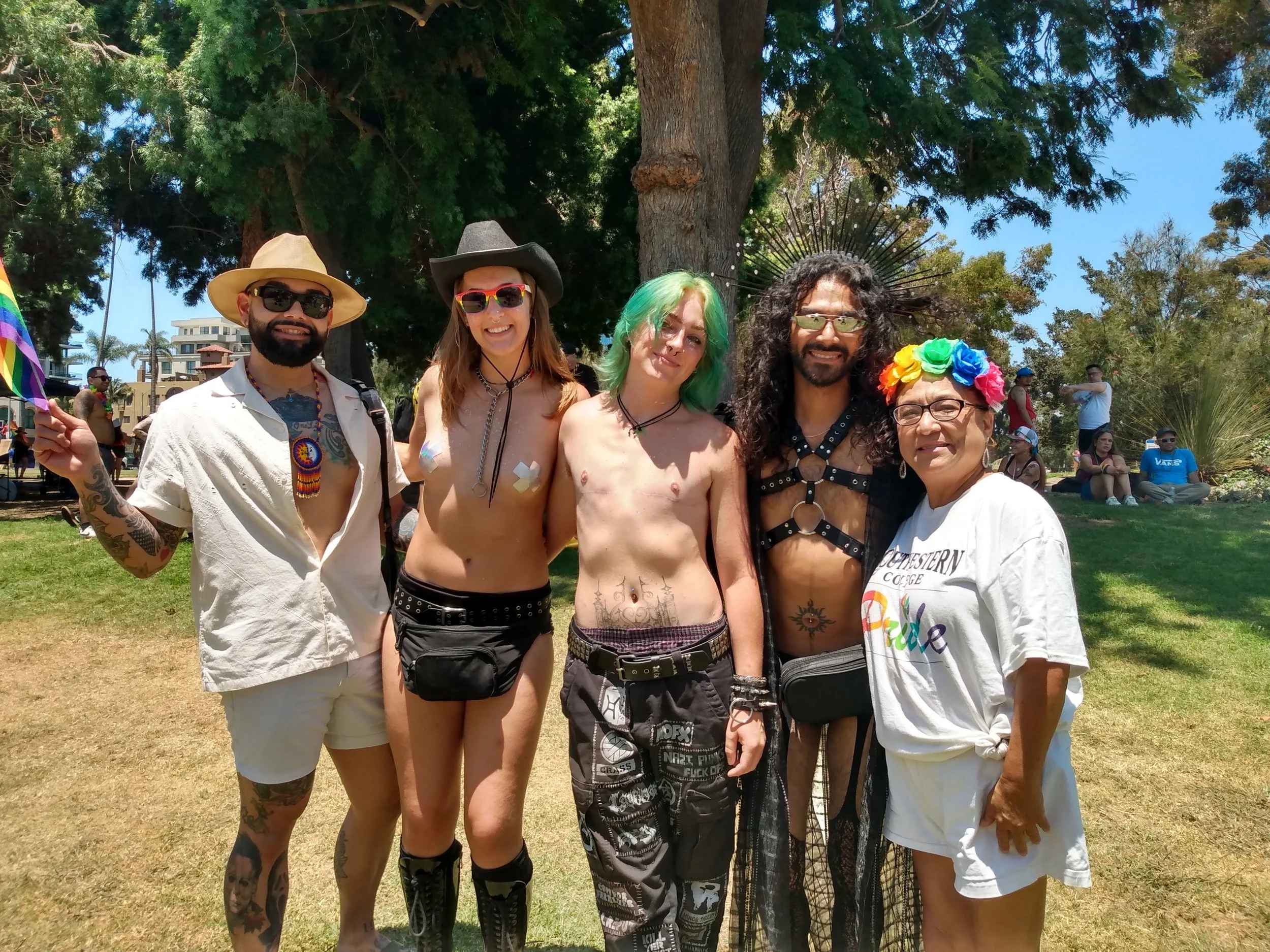 A diverse group of five people is standing outdoors in a park on a sunny day, posing for a photo. They are surrounded by trees, with some sitting on the grass and others standing. The group appears to be celebrating pride or LGBTQ+ pride, with rainbo