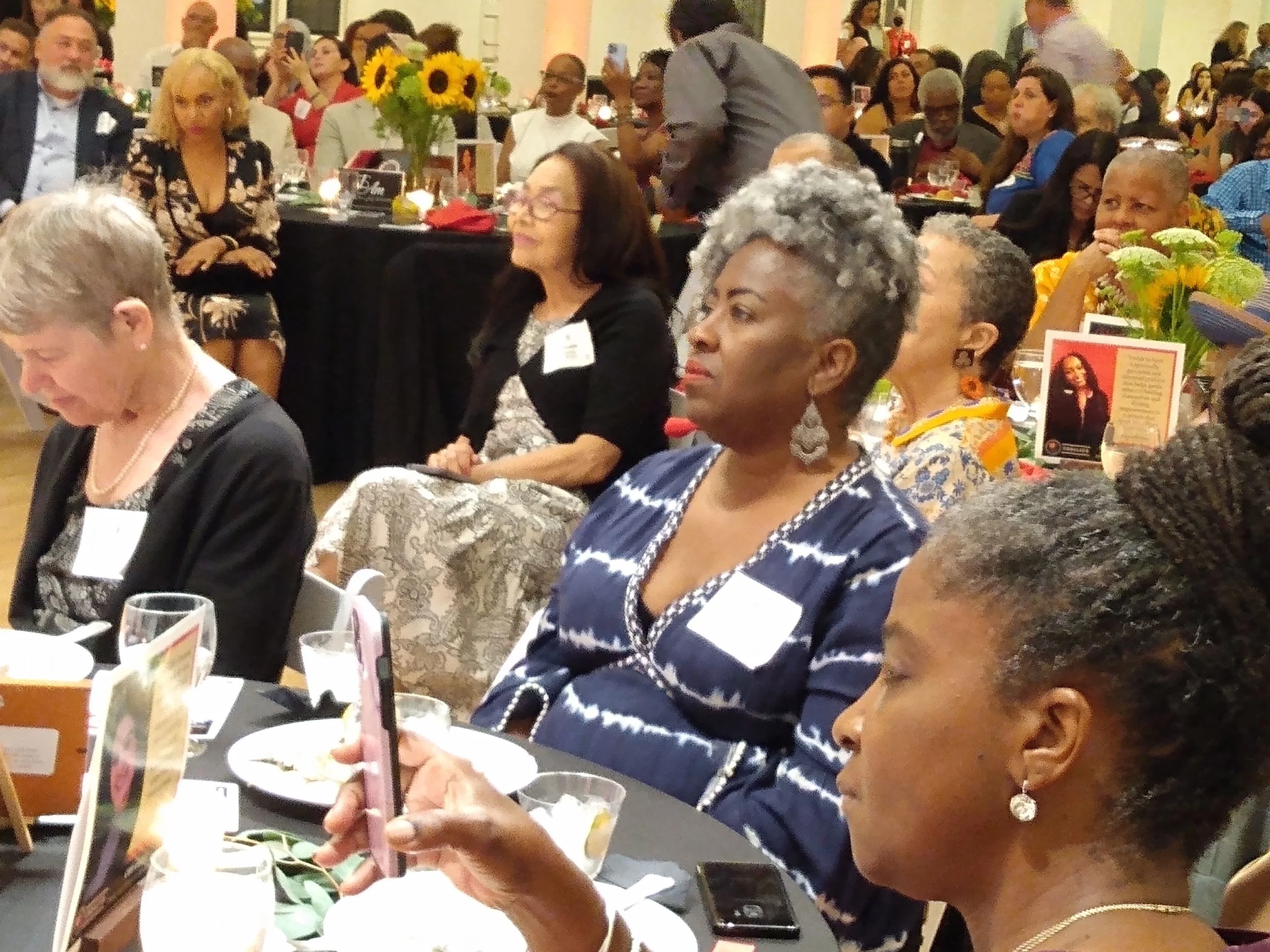 A diverse group of people sitting at round tables during a banquet or dinner event, with some taking photos and others eating. The tables are decorated with flowers, candles, and table cards. The ambiance is lively and festive.