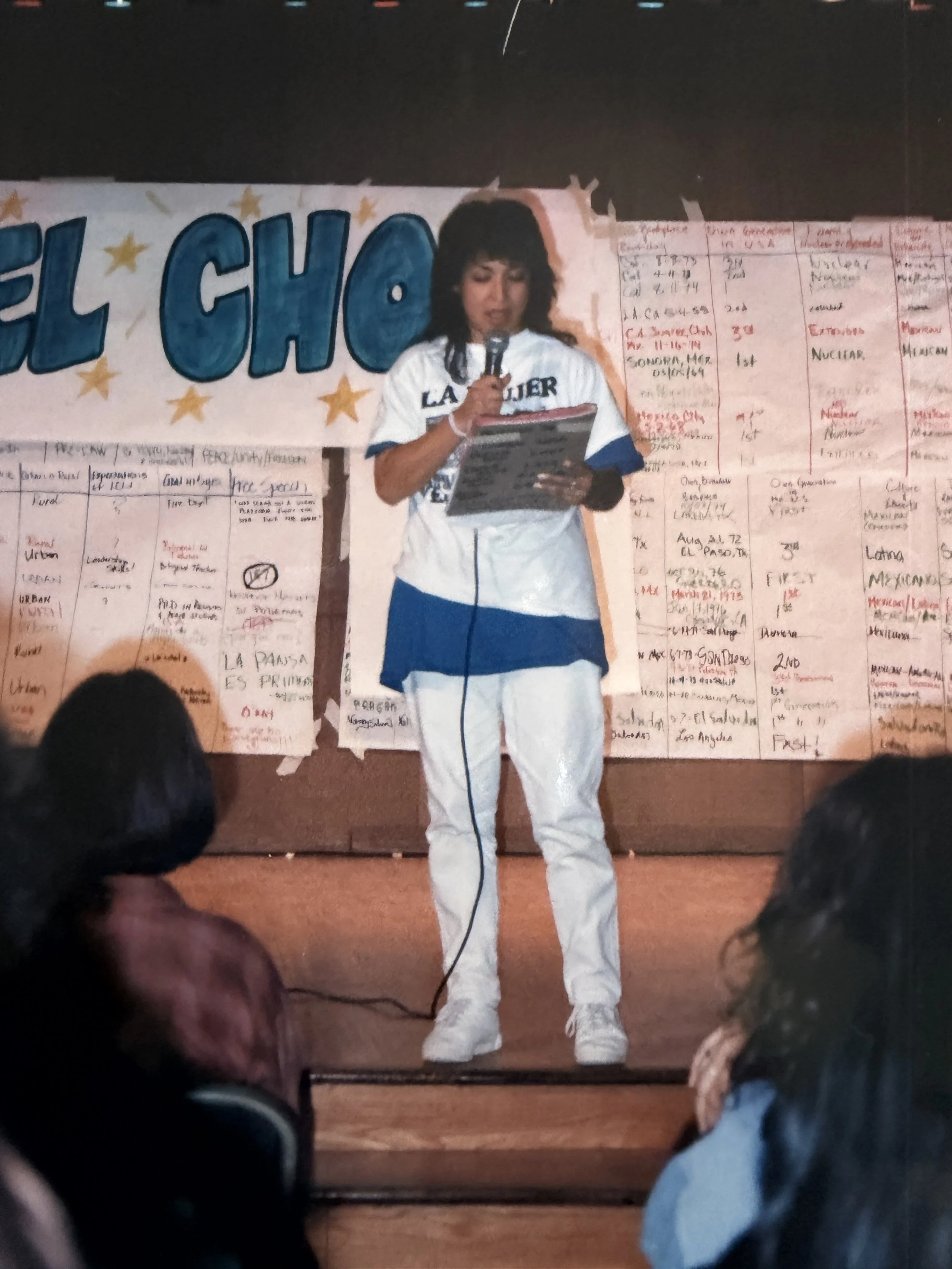 A woman in a white sports uniform speaking into a microphone during a presentation or event. She stands in front of a large banner with the word 'EL CHO' visible, decorated with stars. The background has handwritten notes or charts on whiteboards.