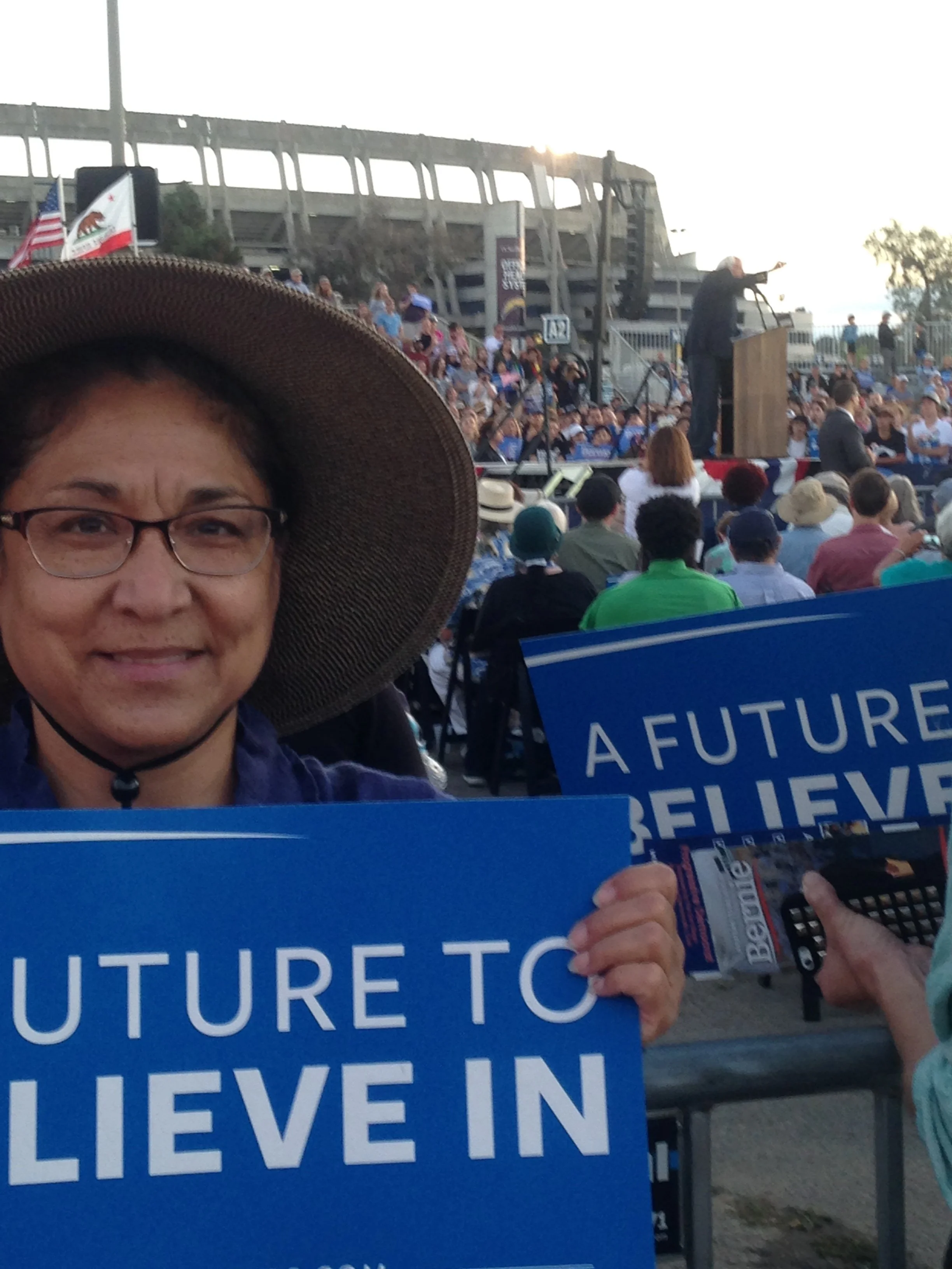 A woman in glasses and a wide-brimmed hat holding a blue sign with white text that reads 'FUTURE TO BELIEVE IN'. In the background, there is a large crowd seated in an outdoor amphitheater, and a speaker at a podium gesturing with his right hand. Fla