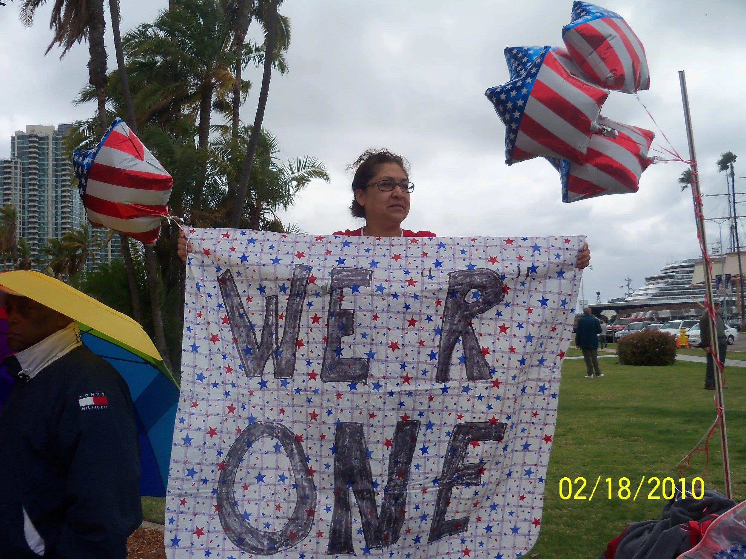 A woman holding a patriotic sign with red, white, and blue star pattern and the word 'VOTE' written on it at an outdoor event. Inflated balloons with American flag design are attached to a pole to her right. Palm trees and high-rise buildings are vis