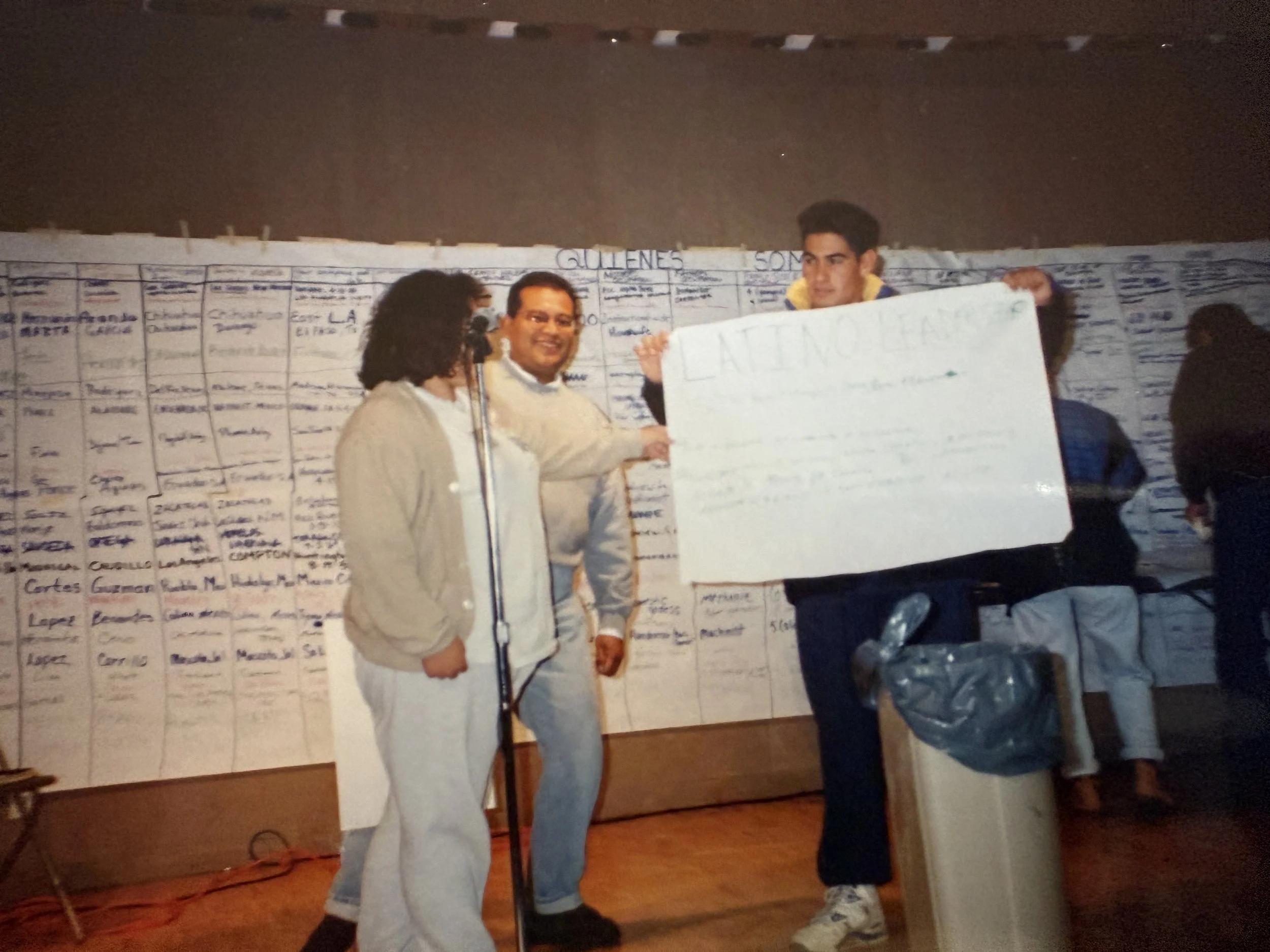 Three people stand in front of a large chart with handwritten information, holding a white poster board, at night.