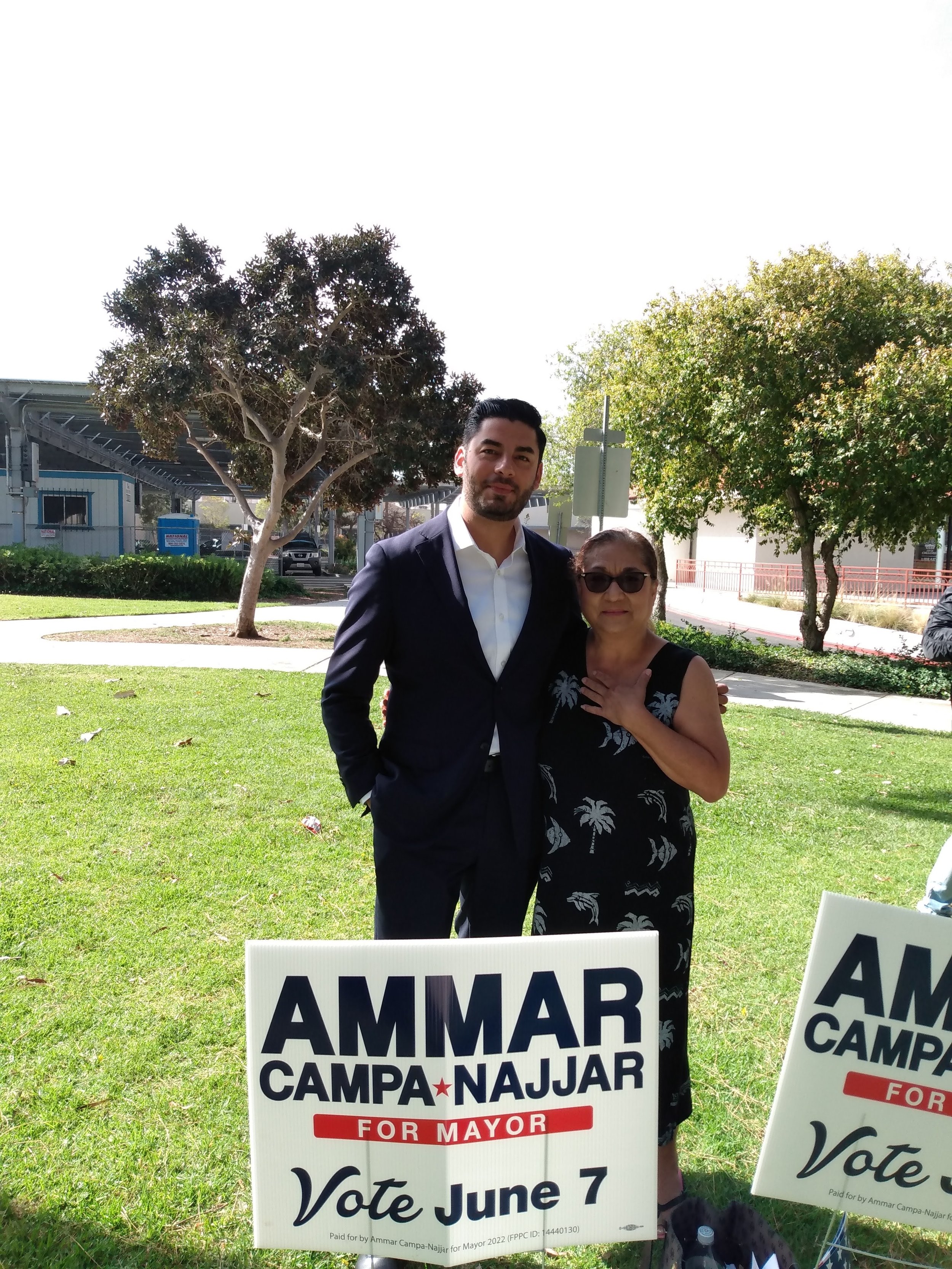 Two people, a man in a suit and a woman wearing sunglasses, standing outdoors behind campaign signs that say "Vote June 7" and "Ammar Campa-Najjar for Mayor." There are trees and a building in the background.