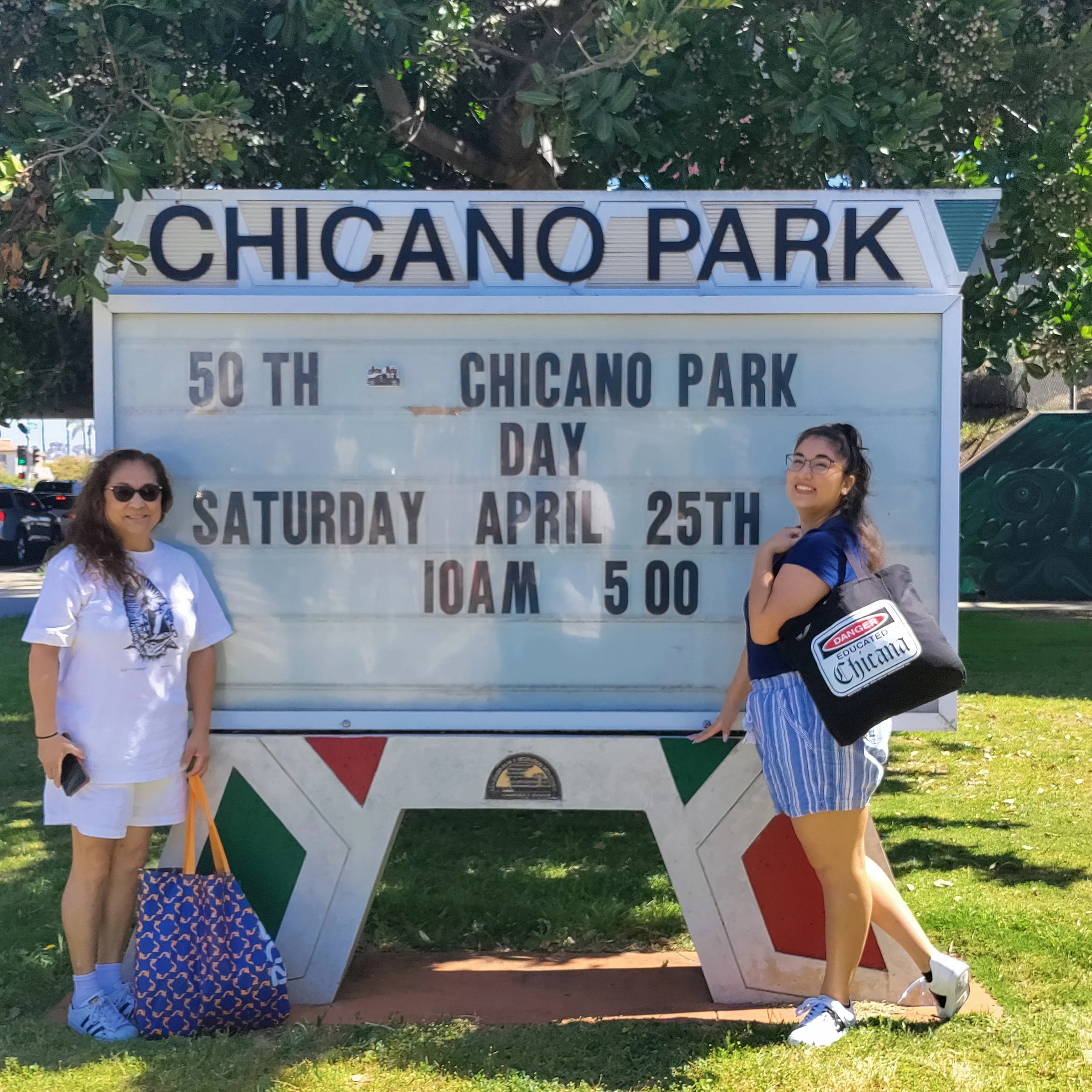 Two women standing in front of a sign for Chicano Park during a celebration for its day. The women are smiling, and the sign indicates the event date is Saturday, April 25th, from 10 a.m. to 5 p.m. The background includes trees and a partial view of 