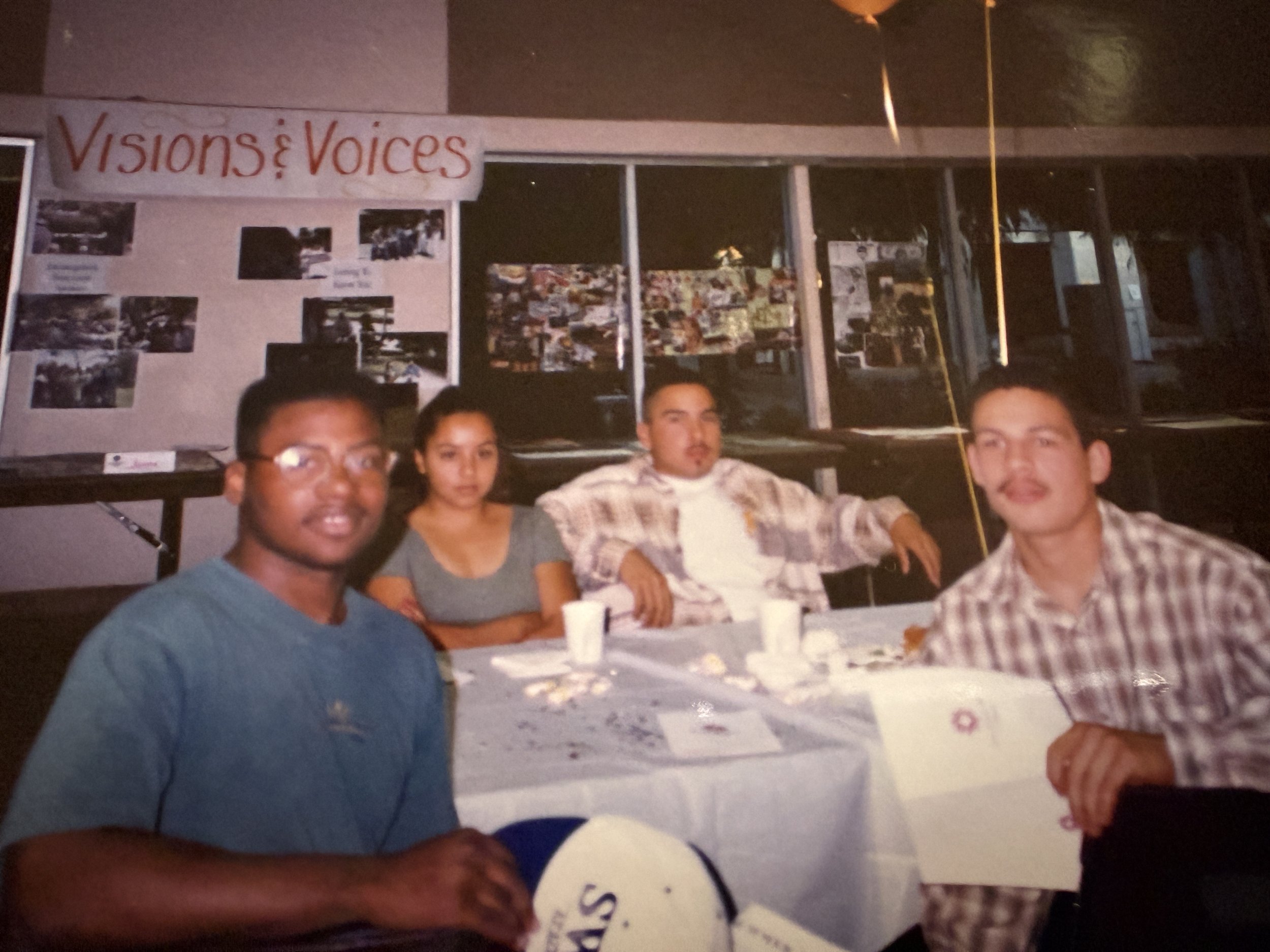 Photo of four young adults seated at a table during an indoor gathering. A large banner with the text 'Visions & Voices' and various photos is visible in the background.