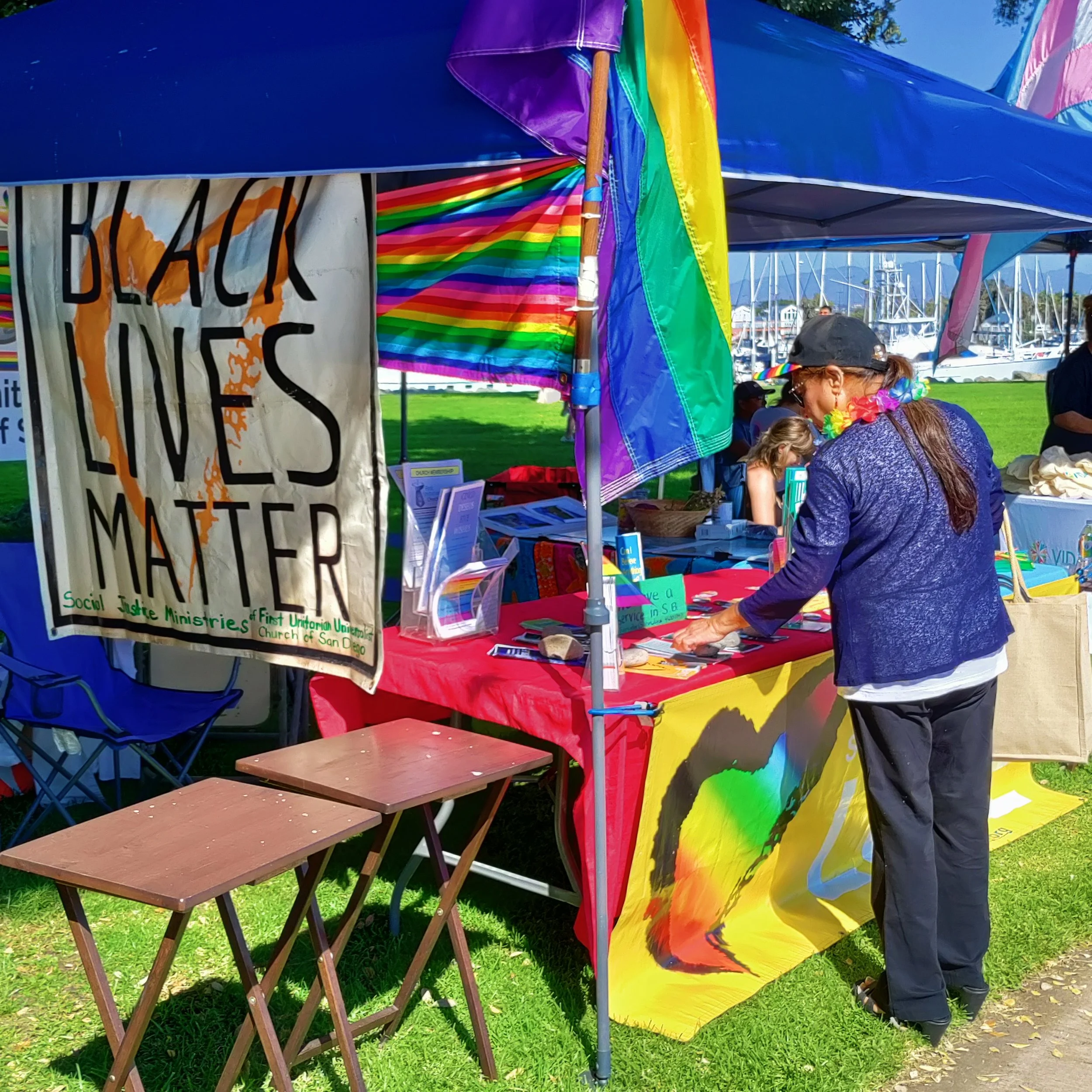 A booth at an outdoor event decorated with rainbow flags, with people gathered around, some wearing rainbow accessories, in a park with boats in the background.