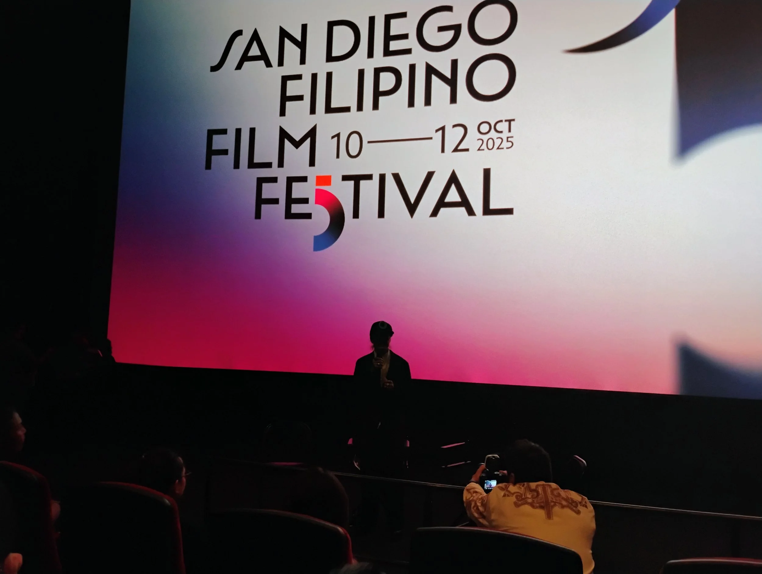 A movie theater auditorium showing a presentation for the San Diego Filipino Film Festival, October 10th to 12th, 2025, with a large screen and silhouettes of audience members in the dark foreground.