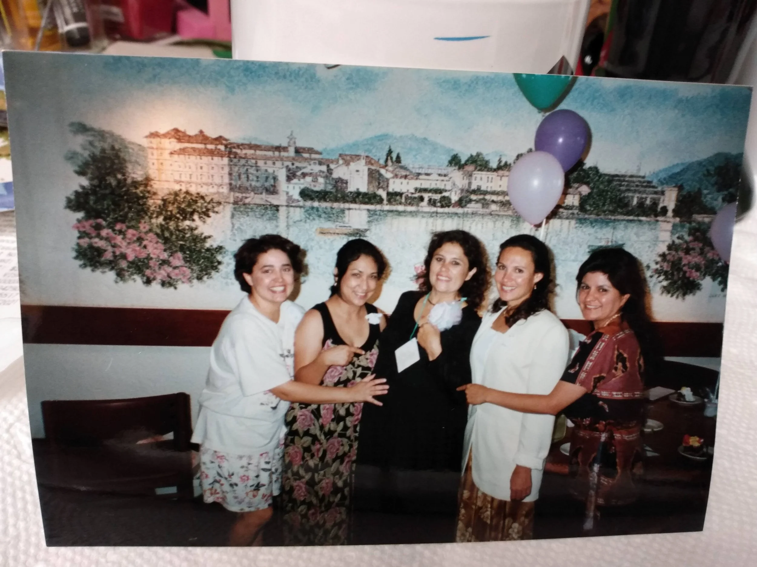 A group of five women standing close together, smiling at the camera, in front of a mural of a European cityscape with historic buildings, water, and mountains in the background. Some women are holding flowers or gifts, and a few are wearing leis or 