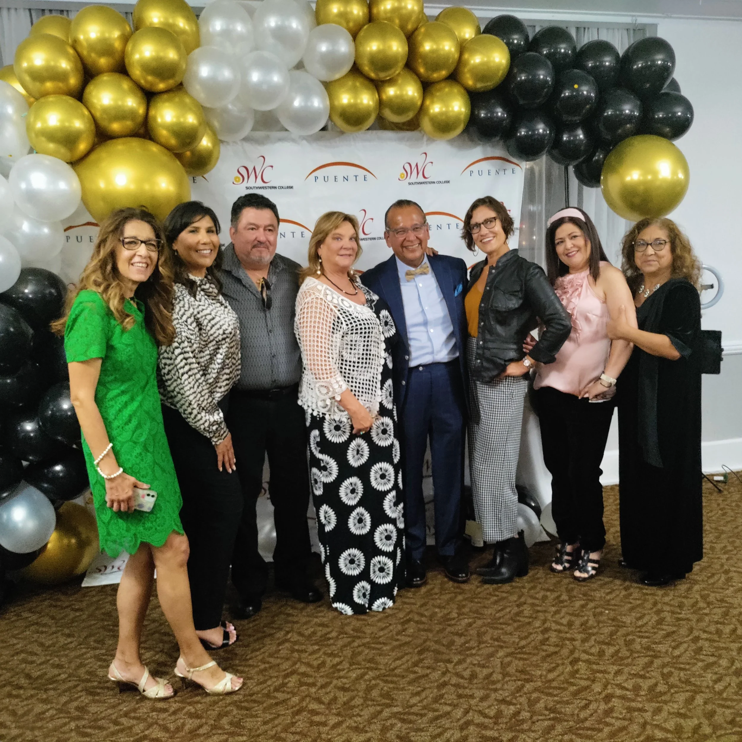 Group of eight adults standing in front of a festive balloon backdrop with swan Lake College and Puente logos, dressed in formal and semi-formal attire, smiling at the camera.