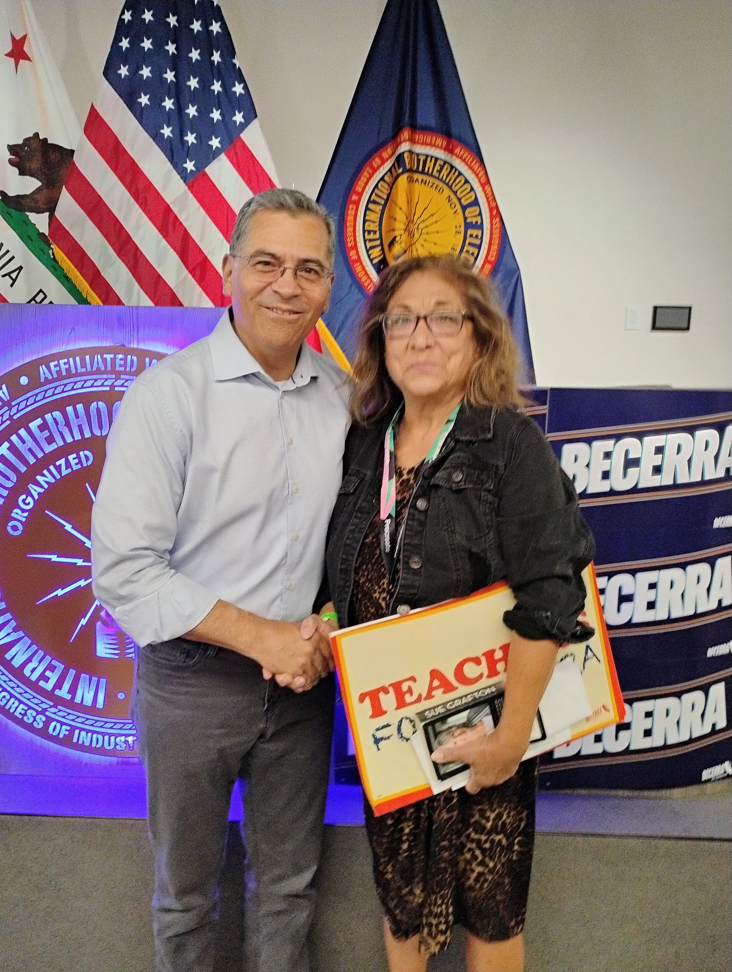 A man and a woman shaking hands at a formal event, standing in front of flags including the American flag and a flag with the emblem of the International Brotherhood of Electrical Workers. The woman is holding a sign that reads 'TEACH' and has other 