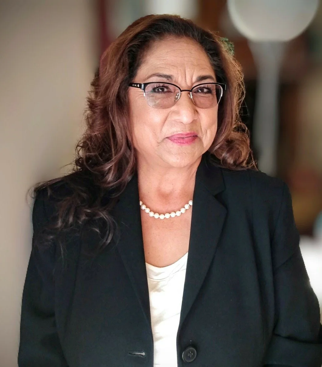 A middle-aged woman with brown curly hair and glasses, wearing a black blazer, a pearl necklace, and a white top, sitting indoors.