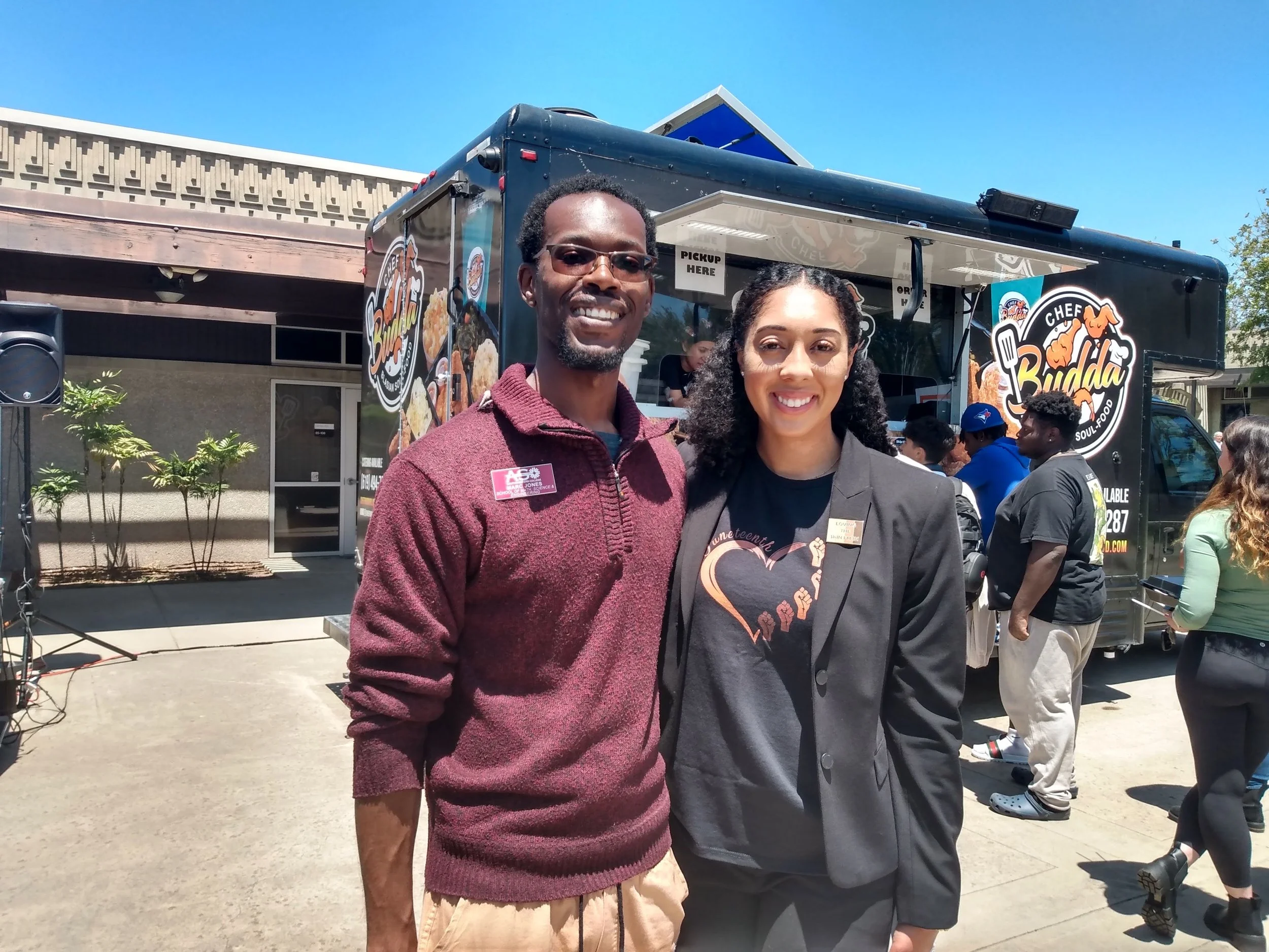 A man and a woman standing close together outdoors in front of a food truck, smiling at the camera during a sunny day.