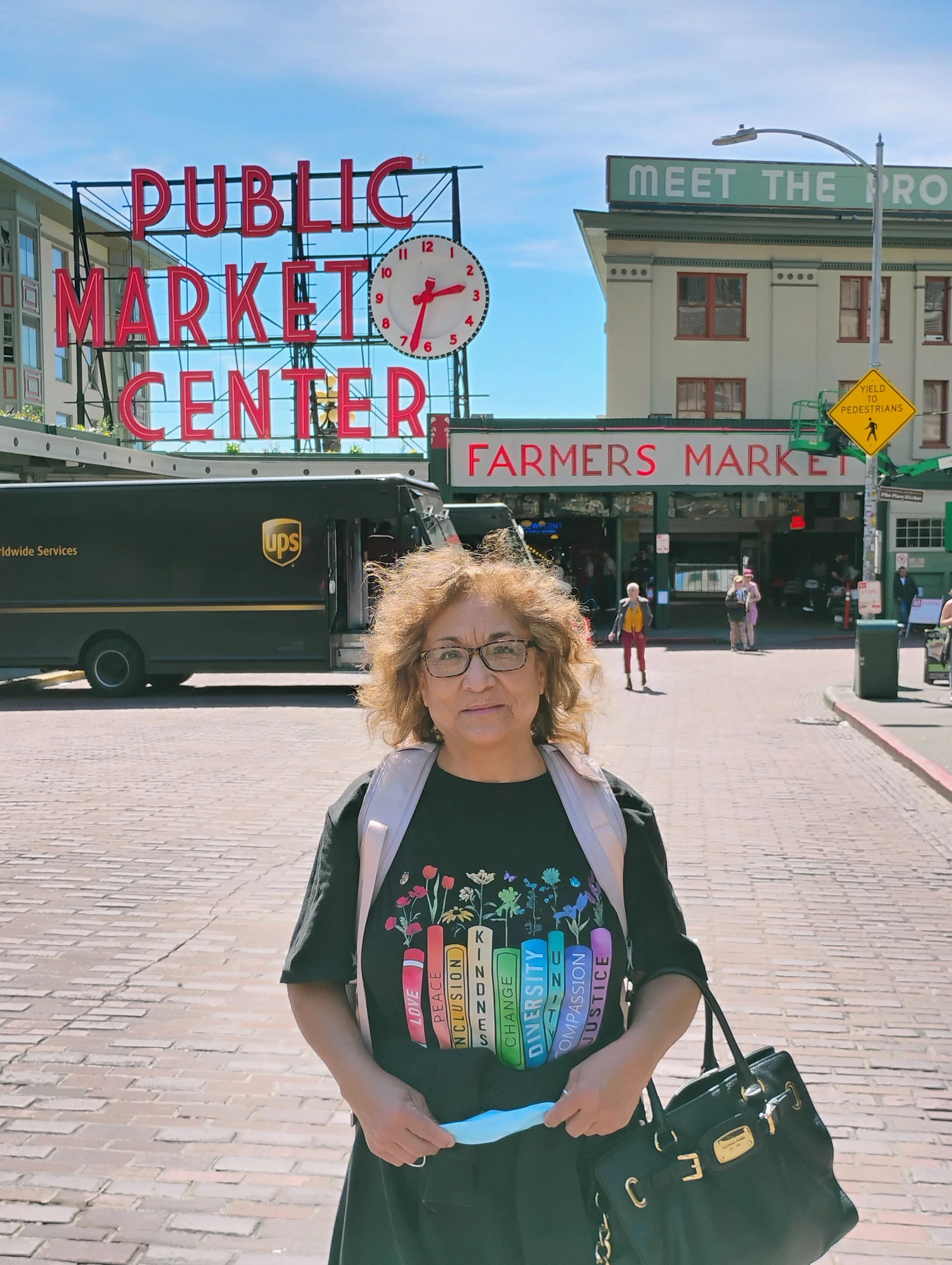 A woman with curly hair and glasses standing outdoors in front of a public market sign and a clock, wearing a black T-shirt with colorful words and holding a black purse.