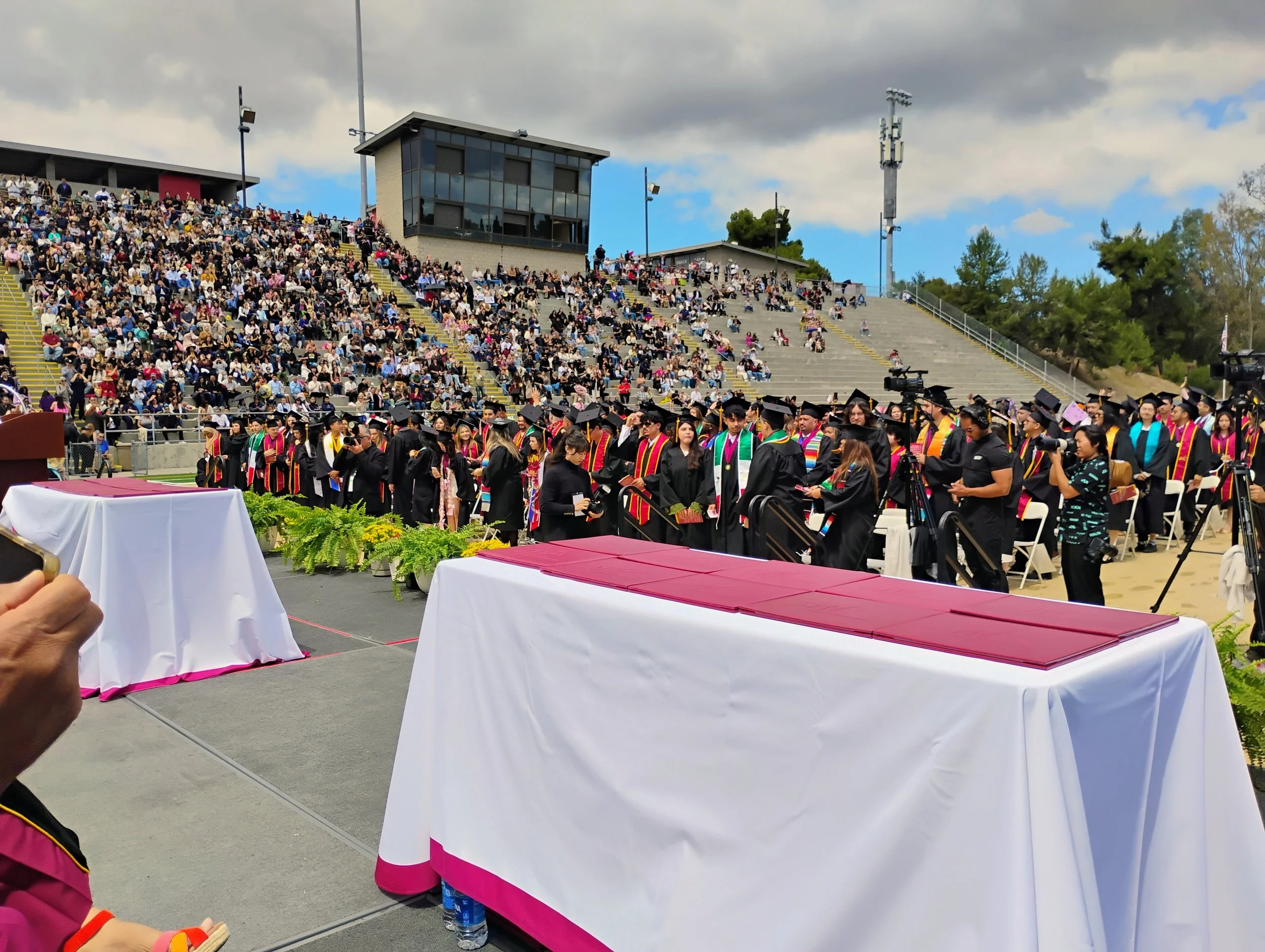 View of a graduation ceremony at an outdoor stadium, with graduates in caps and gowns, an audience seated on bleachers, and cloudy sky overhead.