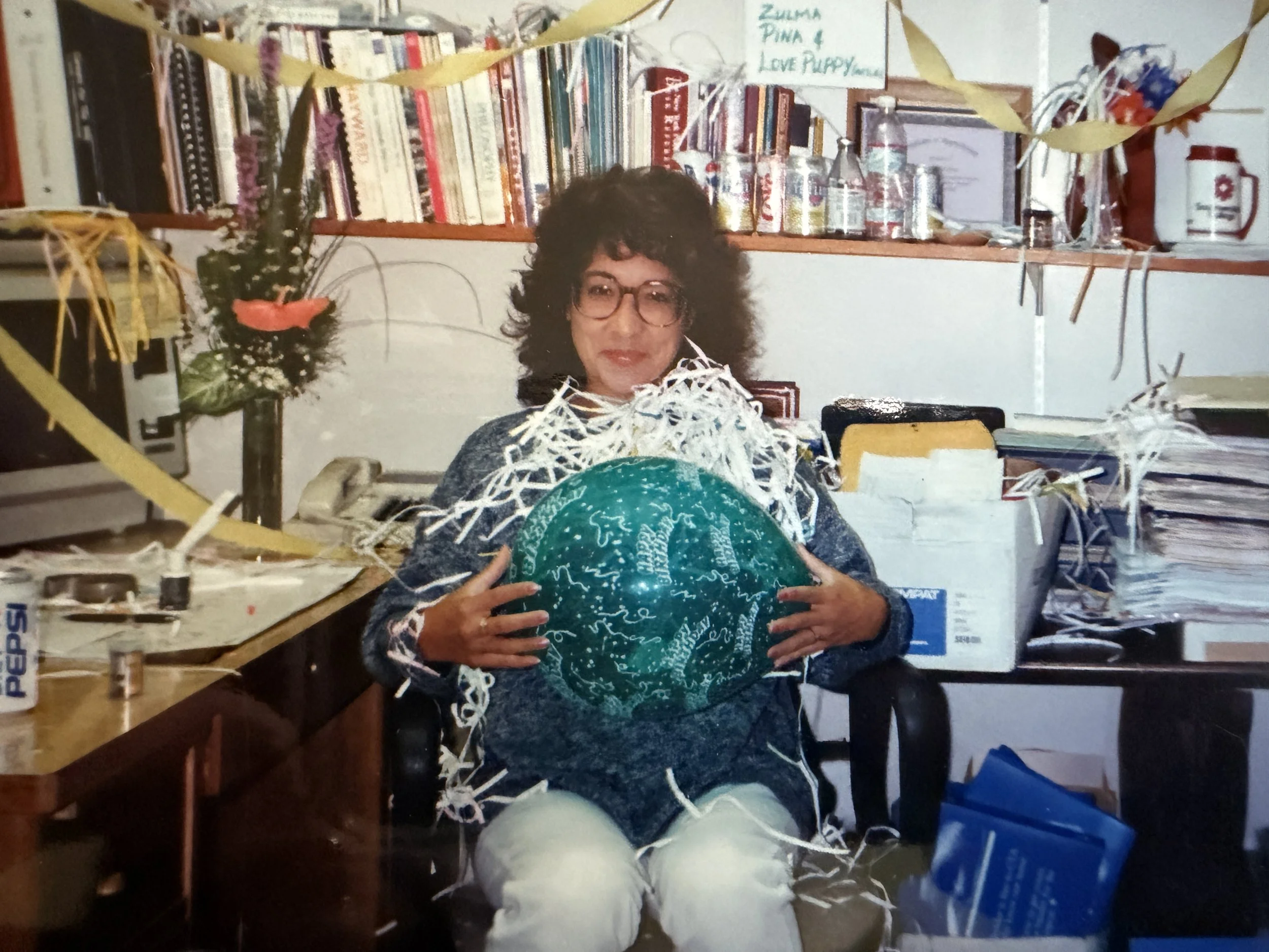 A woman with dark curly hair, glasses, and a cheerful expression is holding a large, green globe in front of her. The background is filled with cluttered shelves, books, bottles, and various objects, indicating a room or office space. There are decor