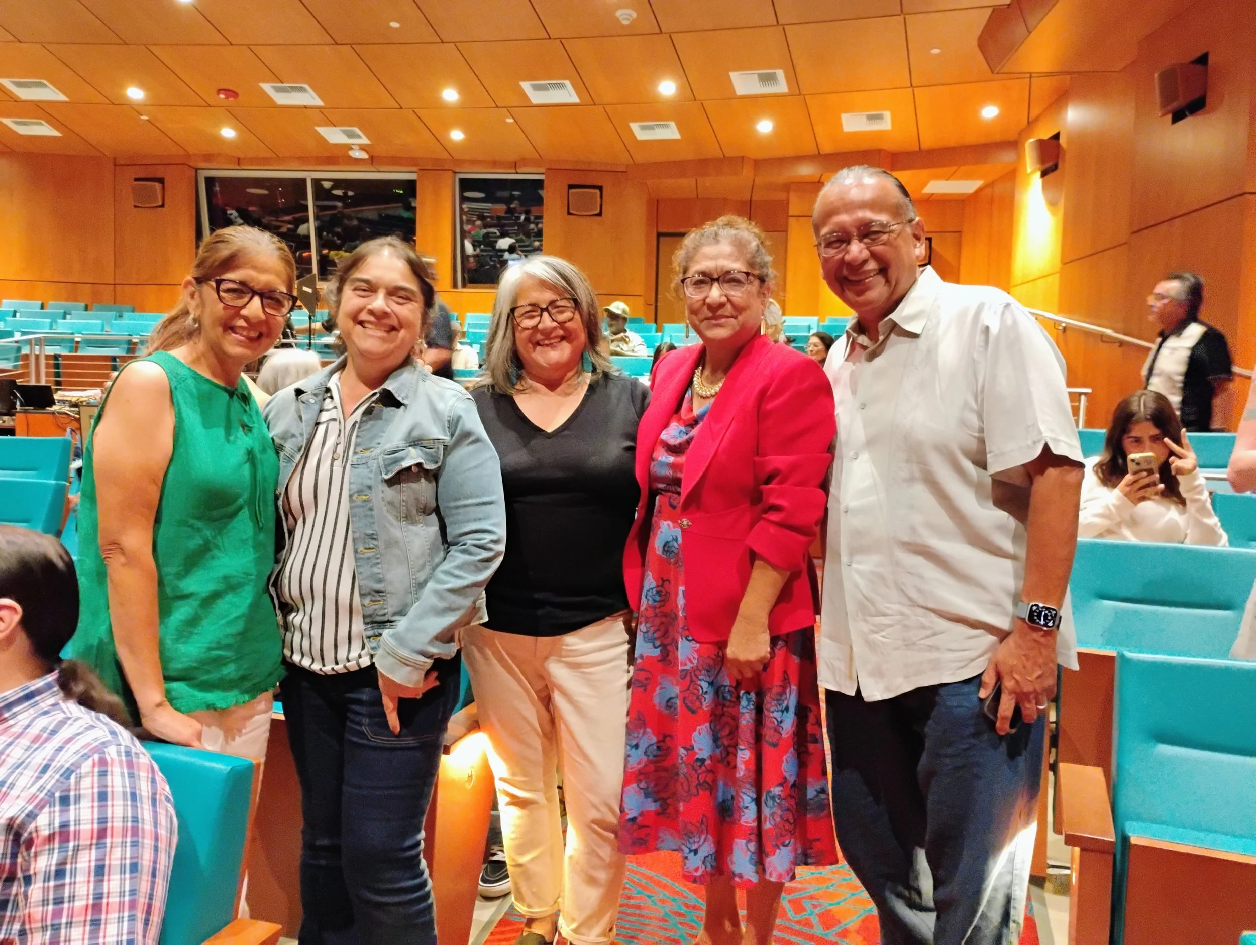 A group of five people standing together inside a theater, smiling at the camera. The background shows teal seats, wooden walls, and large windows with a view of the auditorium.