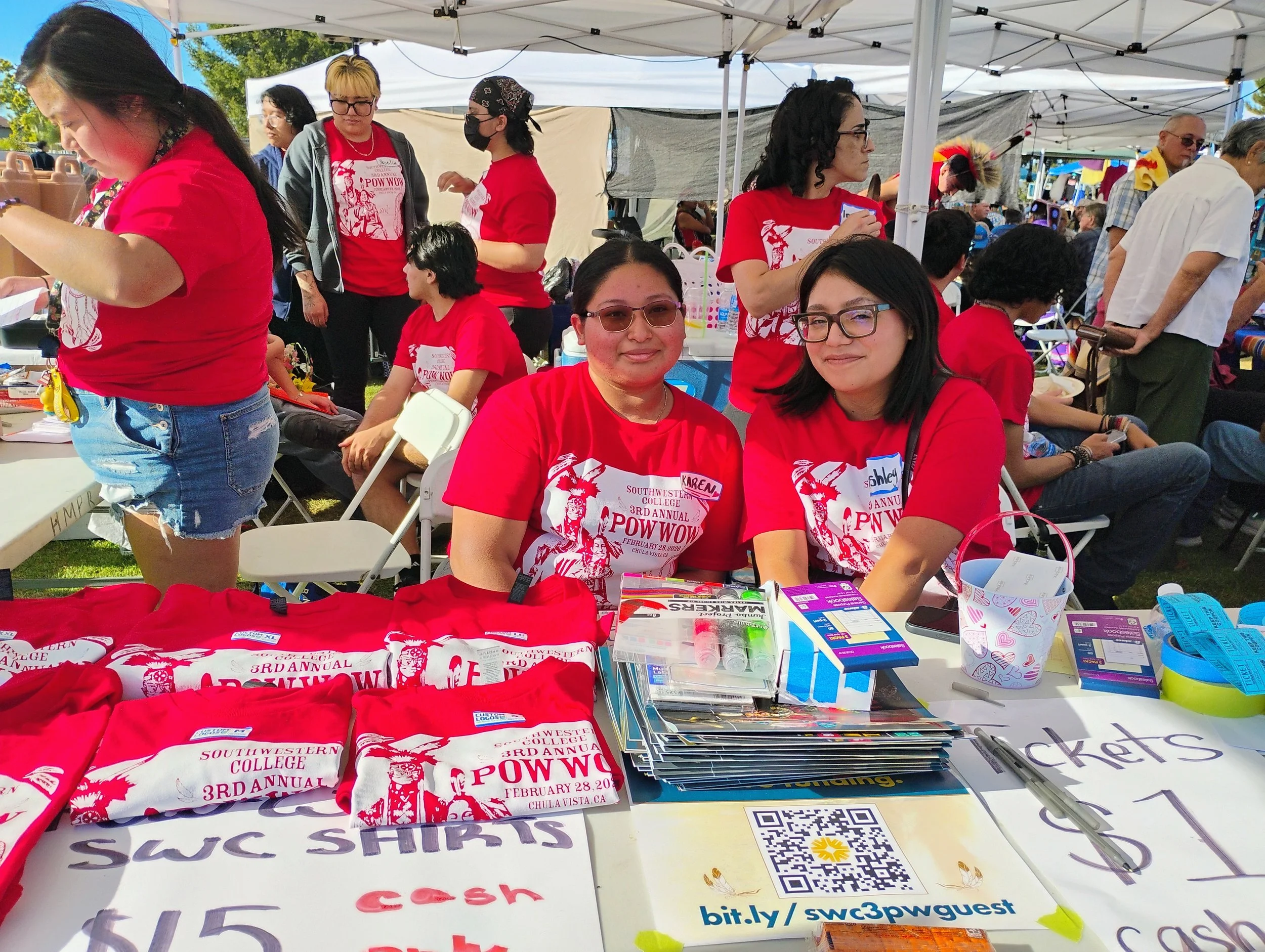 A group of people at an outdoor event under white tents. Some individuals are seated at a table with various items including T-shirts, school supplies, and QR codes. Several people are wearing red shirts with white logos, and the background shows mor