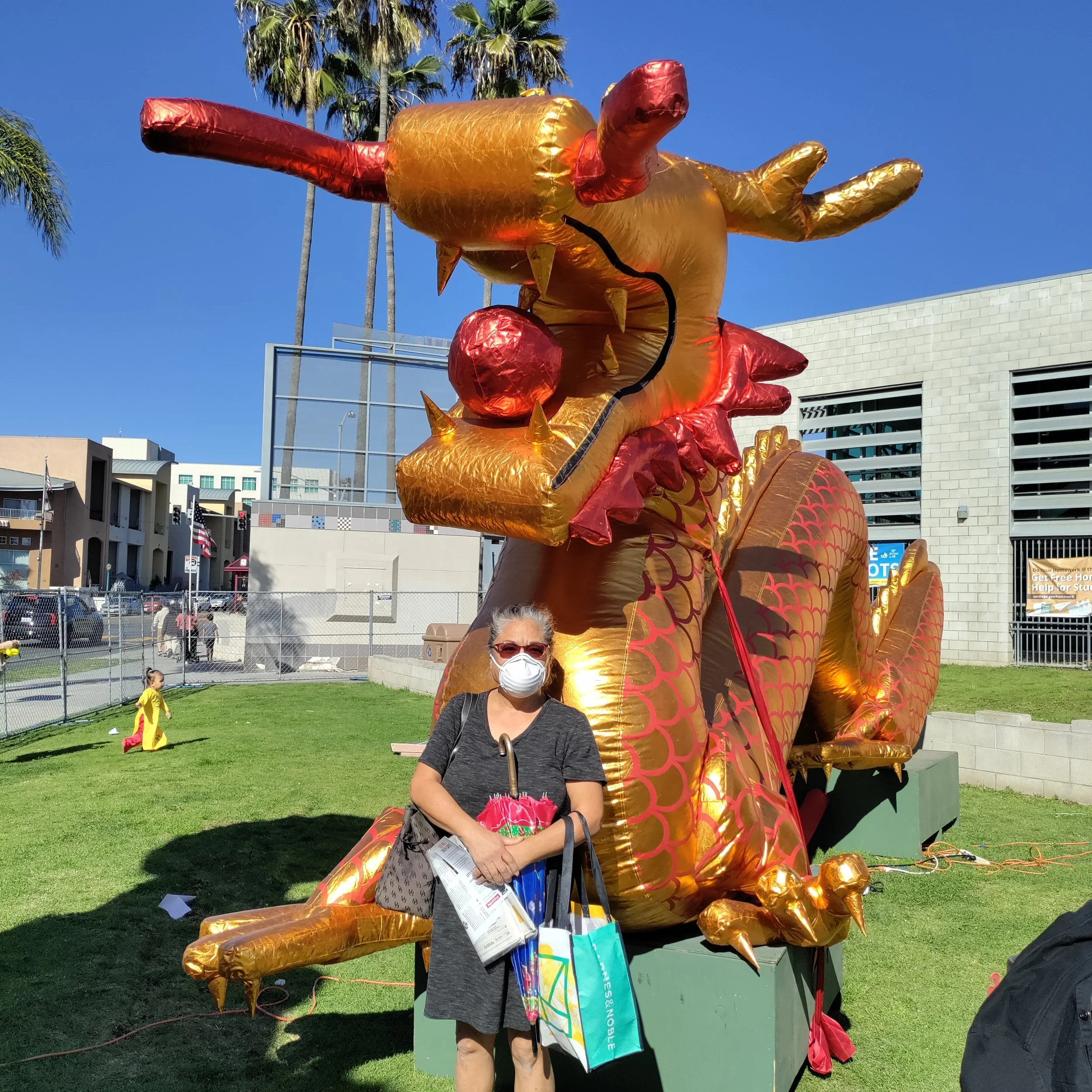 A woman wearing a white face mask and glasses stands in front of a large, gold and red inflatable dragon sculpture at an outdoor event. She is holding shopping bags and pamphlets. The background shows a city street with buildings, palm trees, and a c