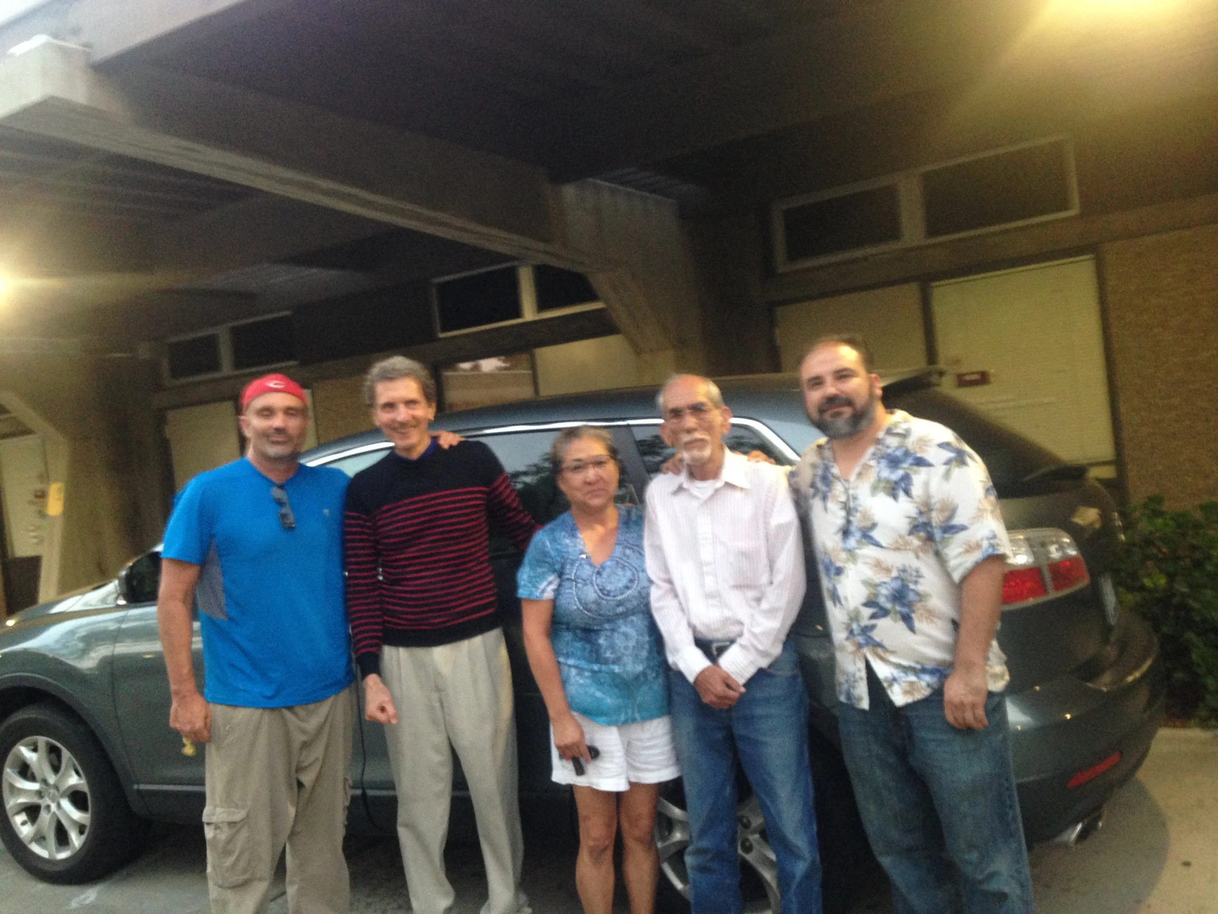 A group of six people, three men and one woman, standing together outside at night in front of a black vehicle, with a building and a dimly lit environment in the background.