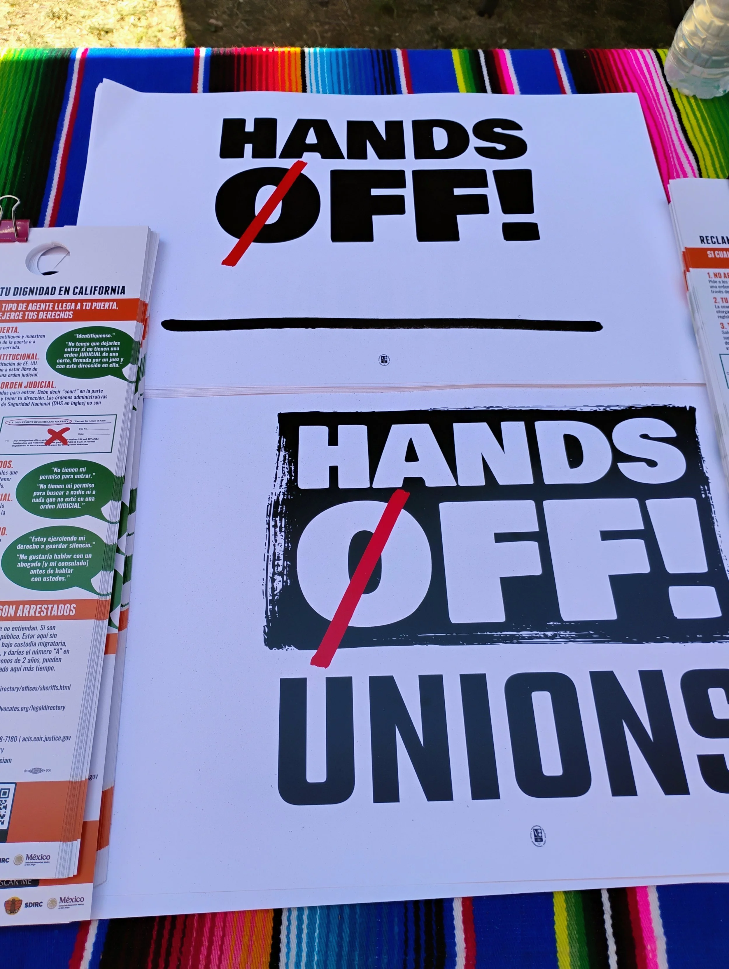 Signs with message 'Hands Off!' placed on a table covered with a colorful, striped cloth, and a partially visible water bottle in the top right corner.