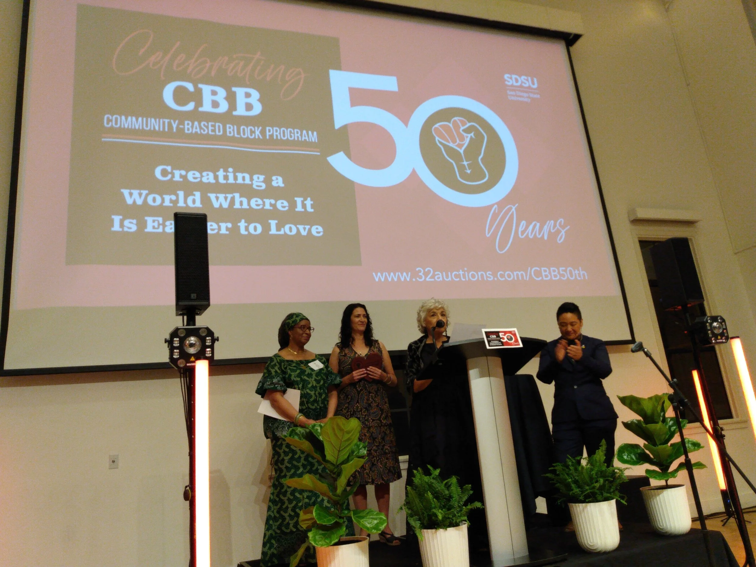 Four women are standing on a stage in front of a large screen displaying the words "Celebrating CBB 50 Years, Creating a World Where It Is Easier to Love." The women are dressed in colorful, patterned dresses and are holding papers. There are potted 