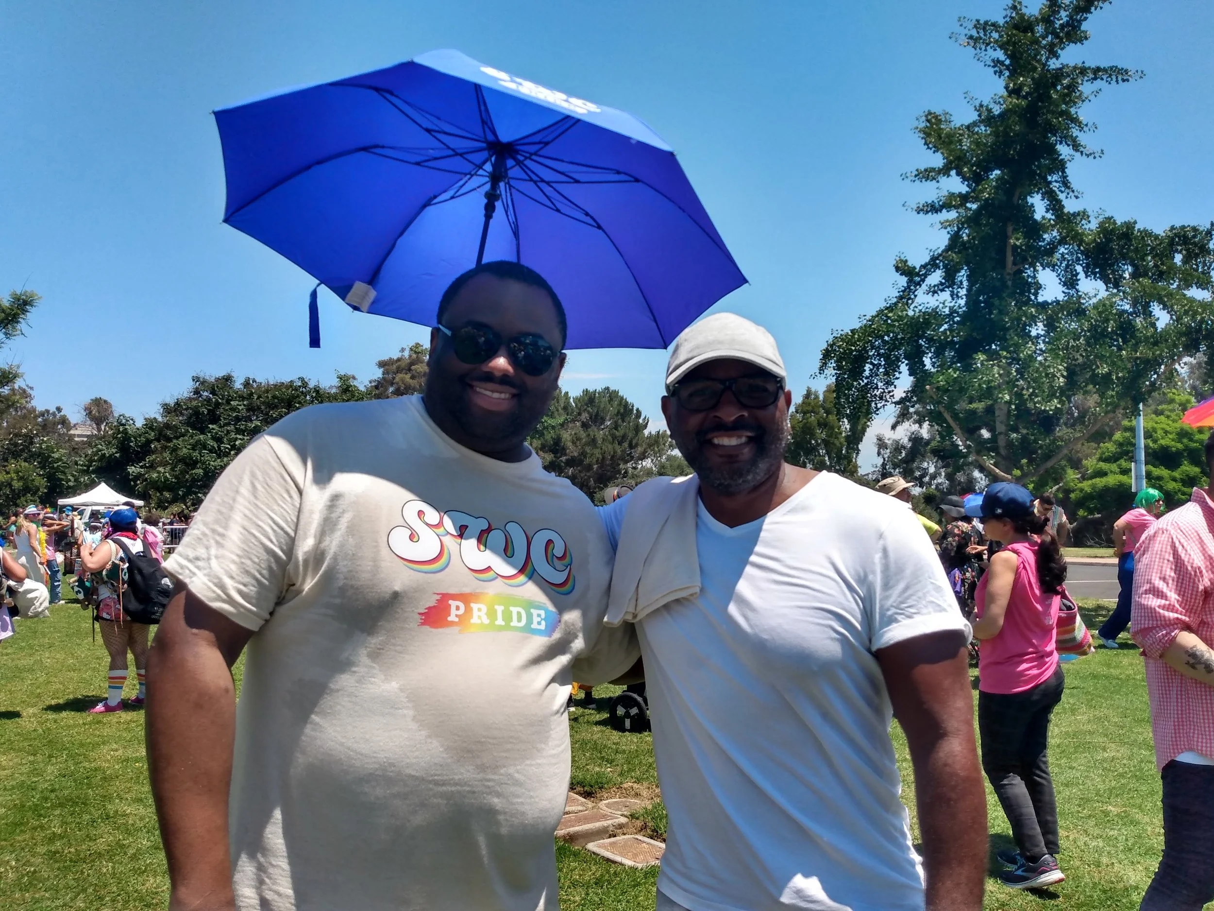 Two men smiling at an outdoor pride event. One is wearing sunglasses and a white T-shirt with rainbow text that says 'Pride,' and the other is wearing a cap and glasses. The man with the Pride T-shirt is holding a large blue umbrella. The background 