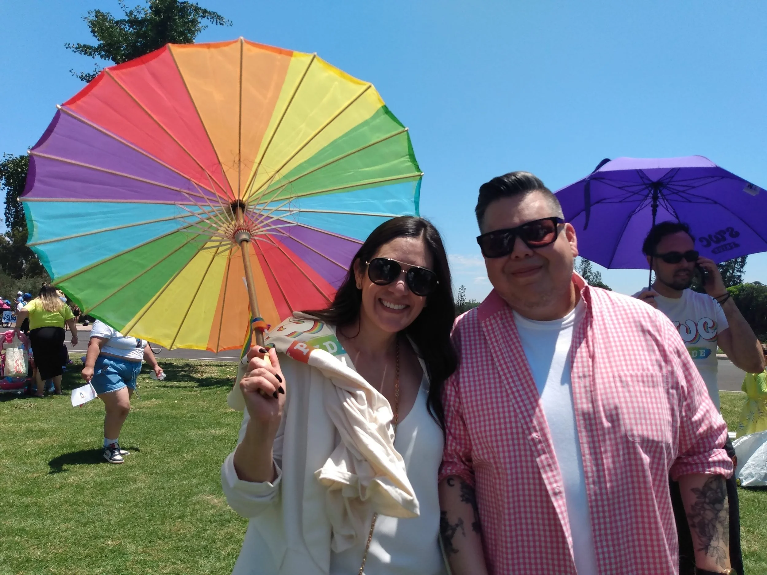 Two people smiling at an outdoor event, holding a large rainbow-colored umbrella. The woman is wearing sunglasses and a white jacket, and the man is wearing sunglasses and a pink checkered shirt. There are other people in the background, some with um