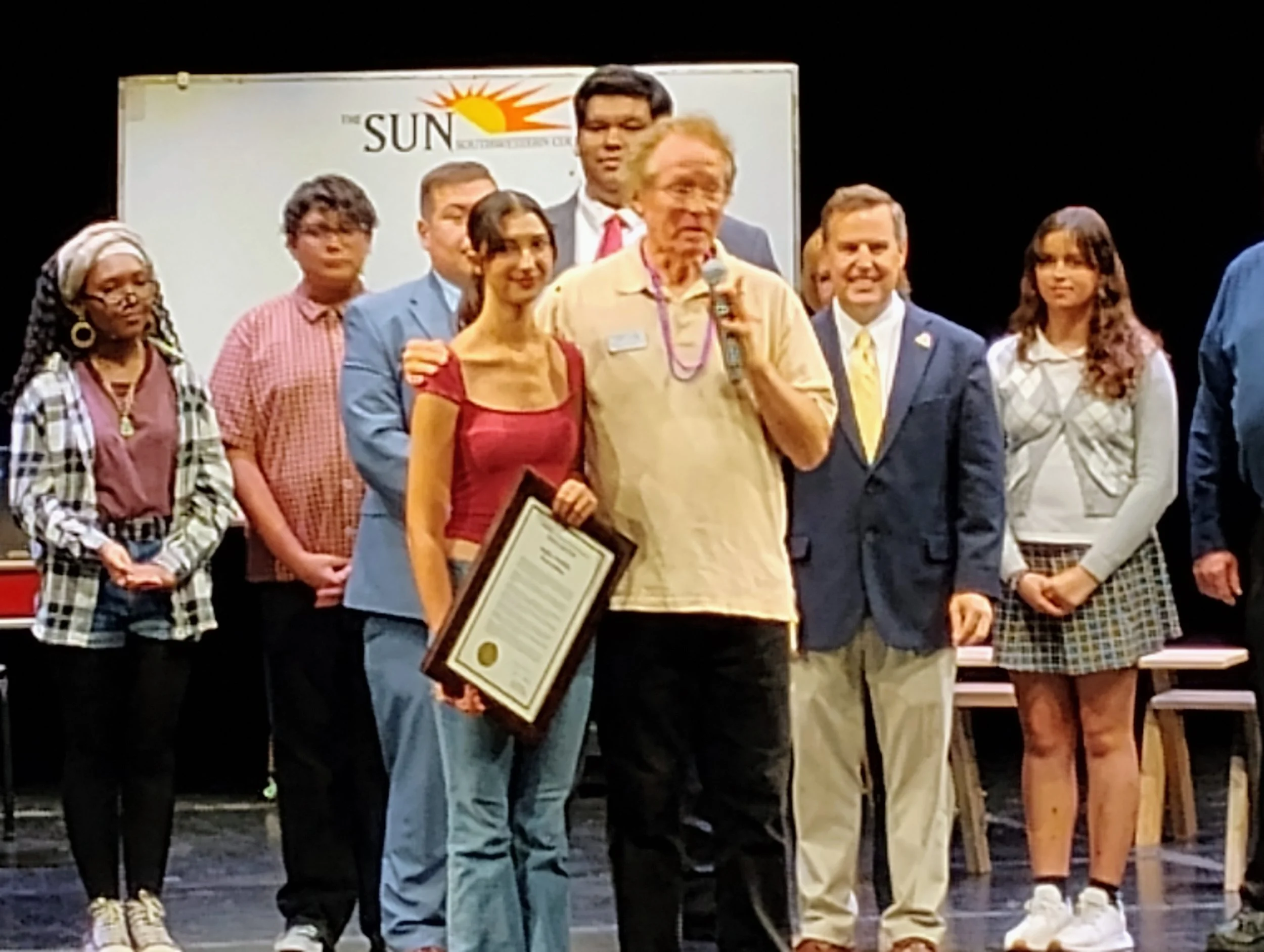 Group of people standing on stage, some holding certificates, in front of a banner that reads 'The SUN' with a sun logo.