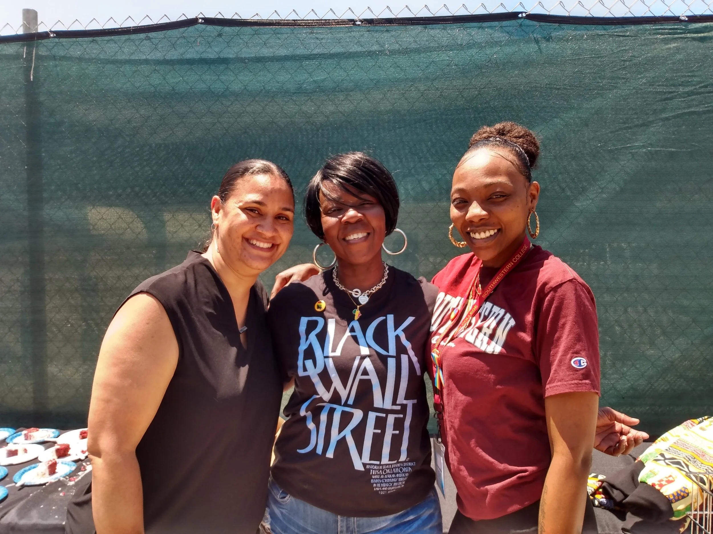 Three women smiling outdoors in front of a green mesh fence. They are wearing casual clothing and have earrings and necklaces. The woman on the right is wearing a maroon shirt with a lanyard around her neck. The woman in the middle has short black ha