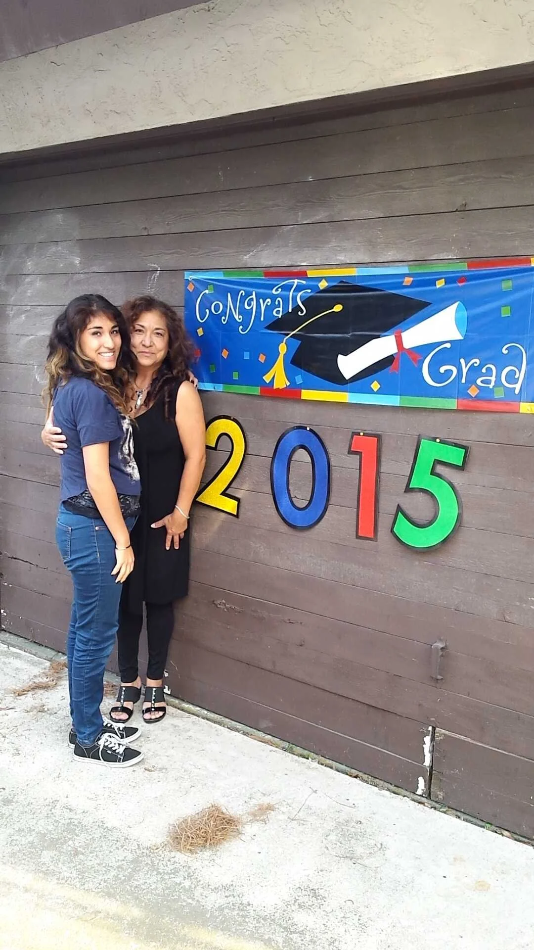 A young woman and an older woman standing together and hugging in front of a graduation banner and colorful year numbers "2015" on a wooden wall.