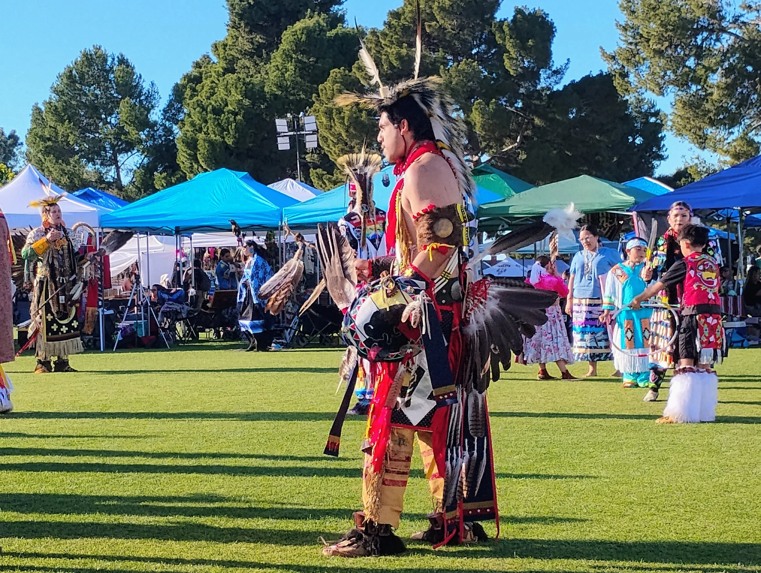 Native American dancers performing at an outdoor cultural event with tents and trees in the background.