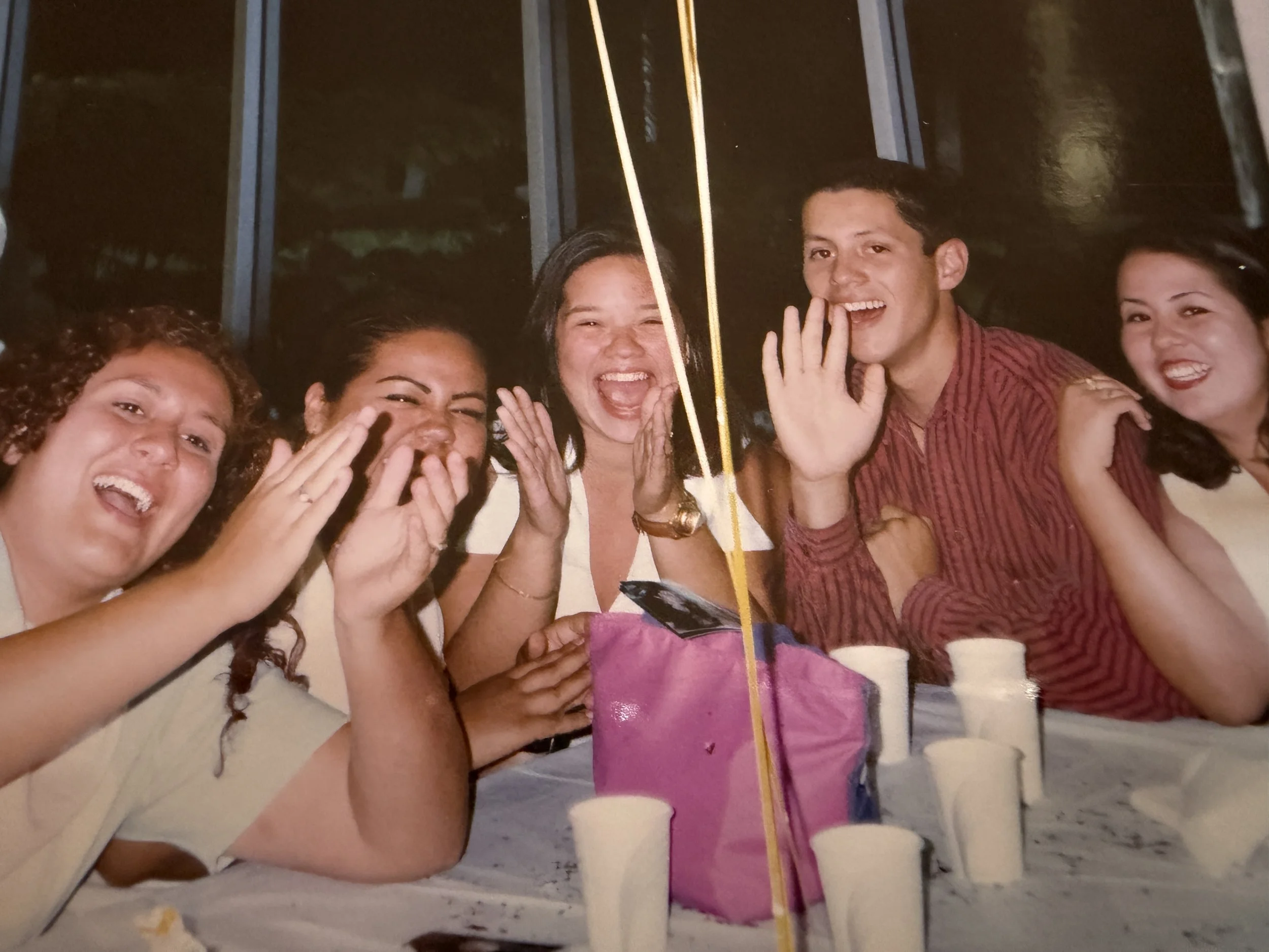 Group of smiling friends at a beach party with a pink birthday cake, balloons, and drinks.