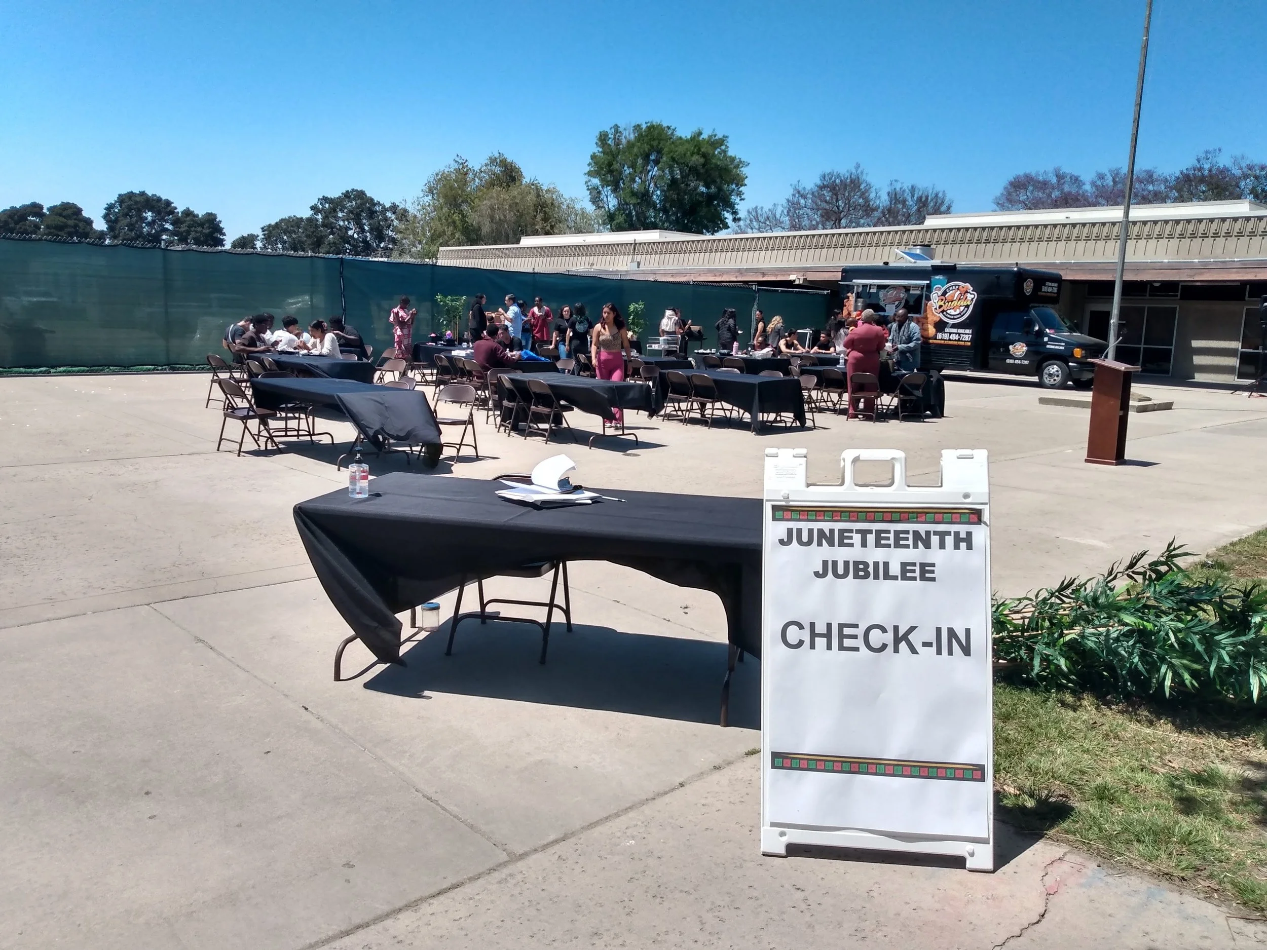 Outdoor scene with tables and chairs set up for an event, food trucks and people gathered in the background, including a sign that reads 'Juneteenth Jubilee Check-In'.
