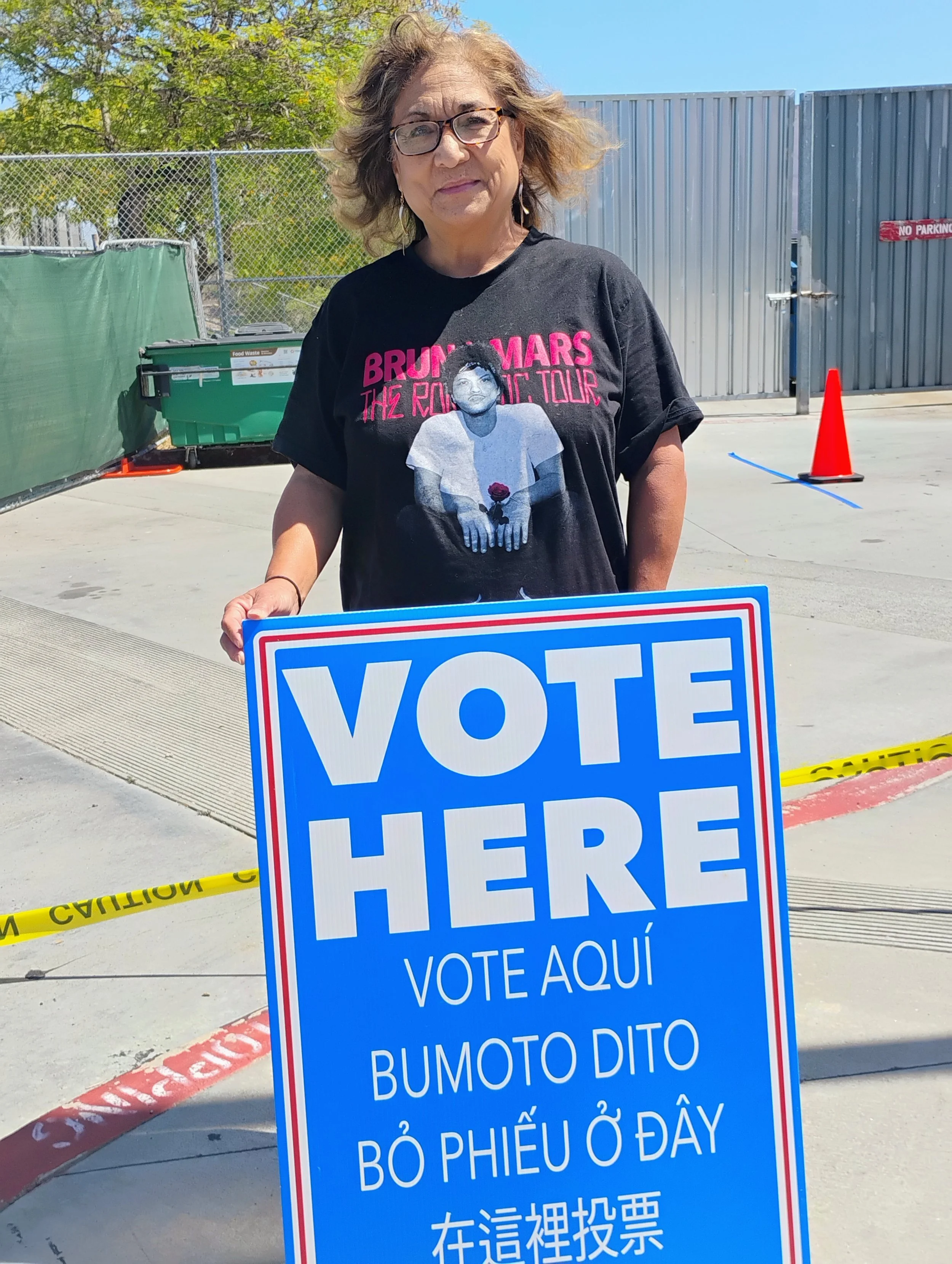 Woman standing outside holding a sign that says 'VOTE HERE' in English and multiple languages including Vietnamese and Chinese. She is wearing glasses and a black t-shirt with a graphic of a man in a white shirt and pink lettering. There are orange traffic cones, yellow caution tape, and a green bin behind her, with a chain-link fence and trees in the background.