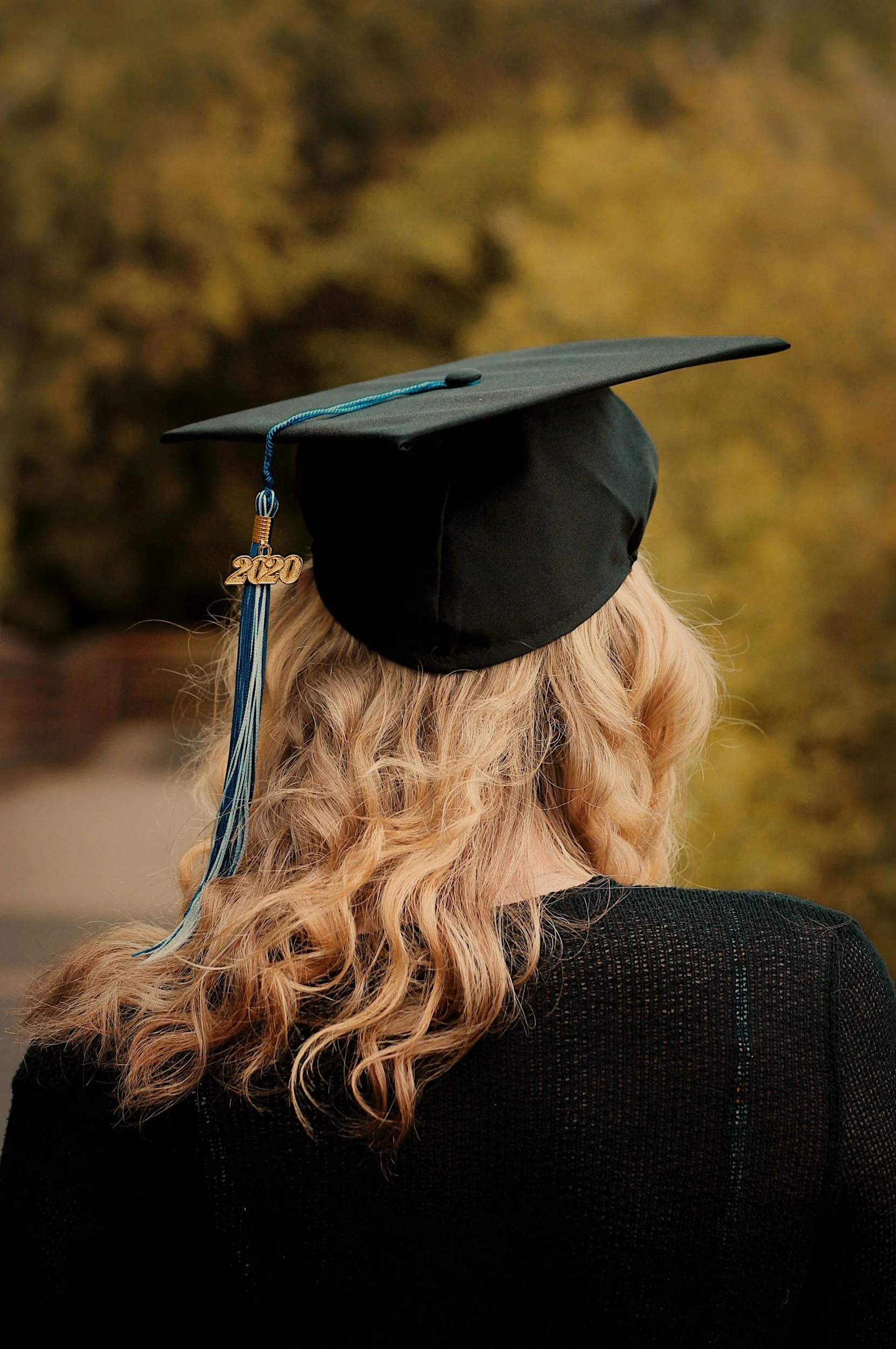 Back of a person wearing a black graduation cap with a blue tassel and a gold charm that reads '2020', with long, curly blonde hair, standing outdoors in a blurred autumn background.