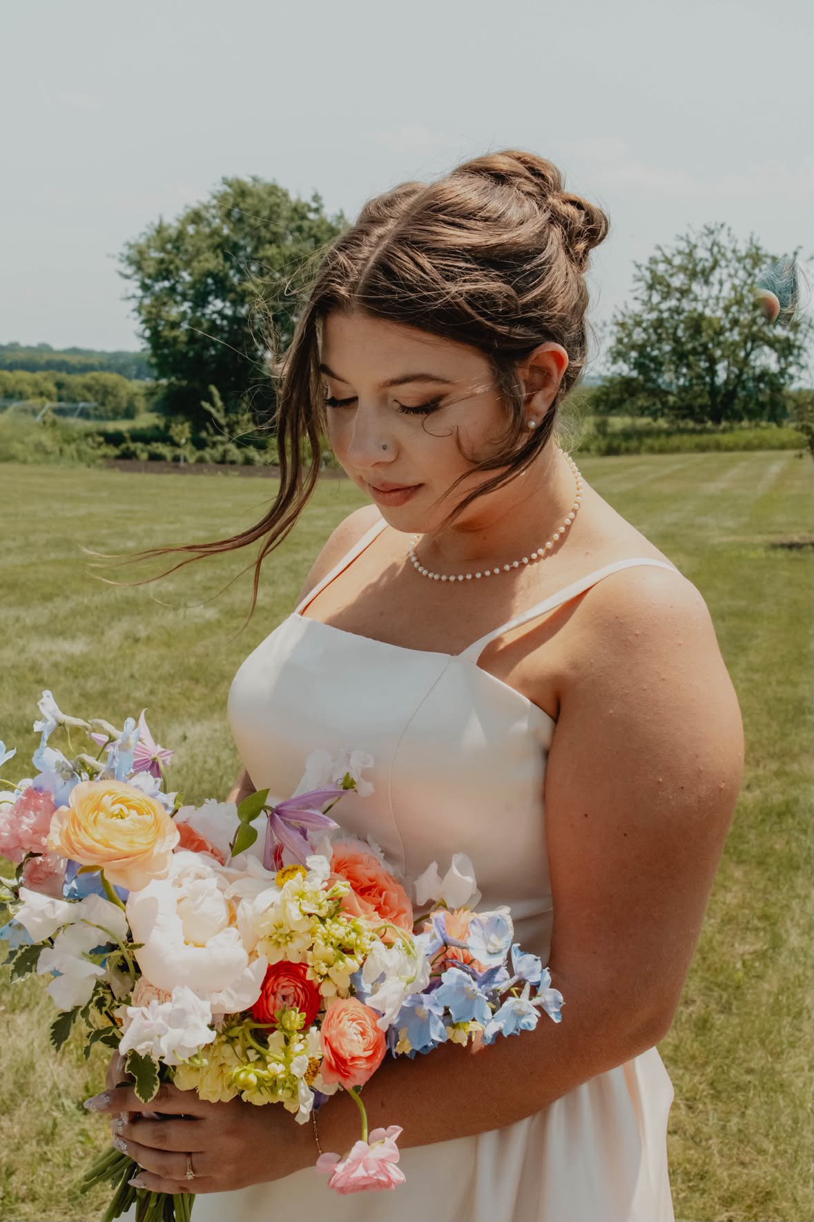 A bride in a white dress and beautiful makeup, holding a large bouquet of colorful flowers, in a grassy field during the day in Madison, WI.