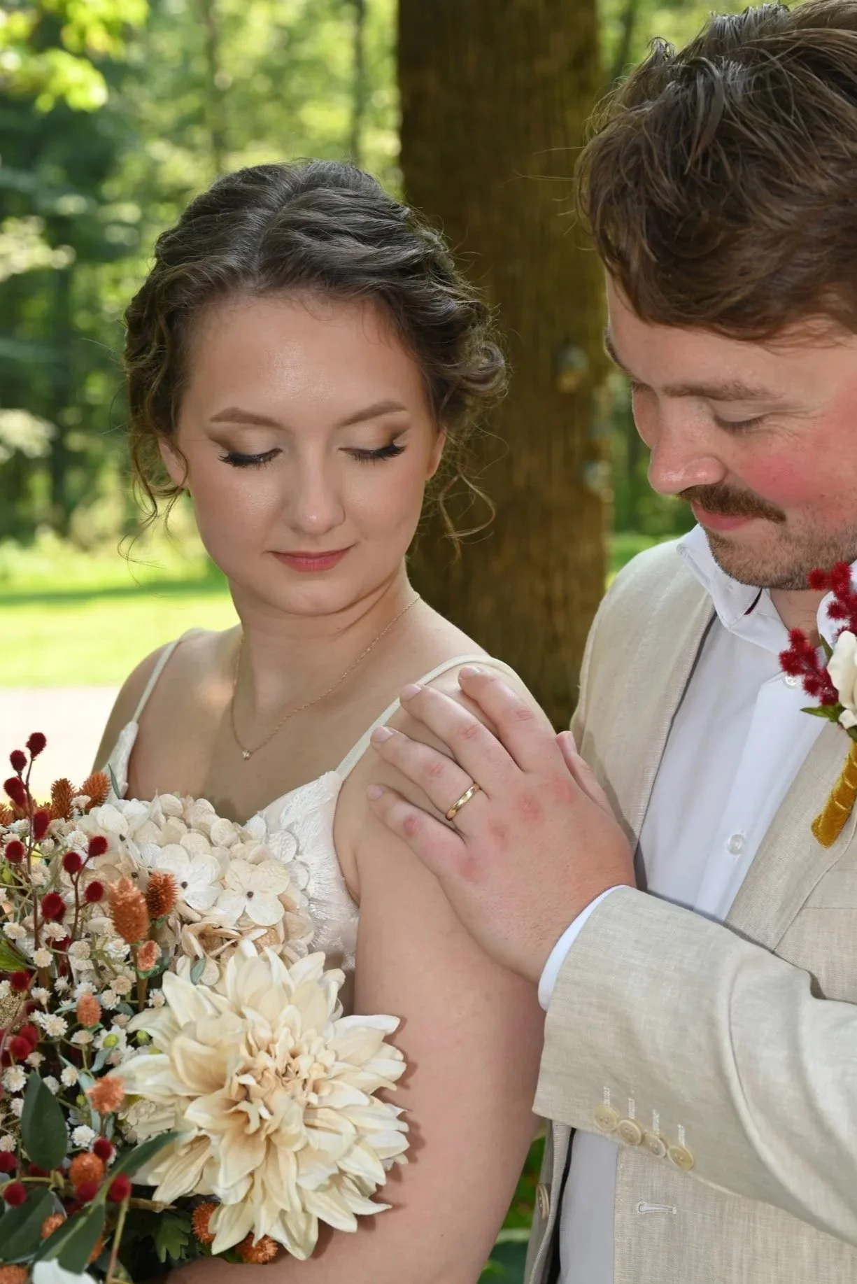 A bride and groom in a garden, with the groom gently touching the bride's shoulder, both smiling softly.