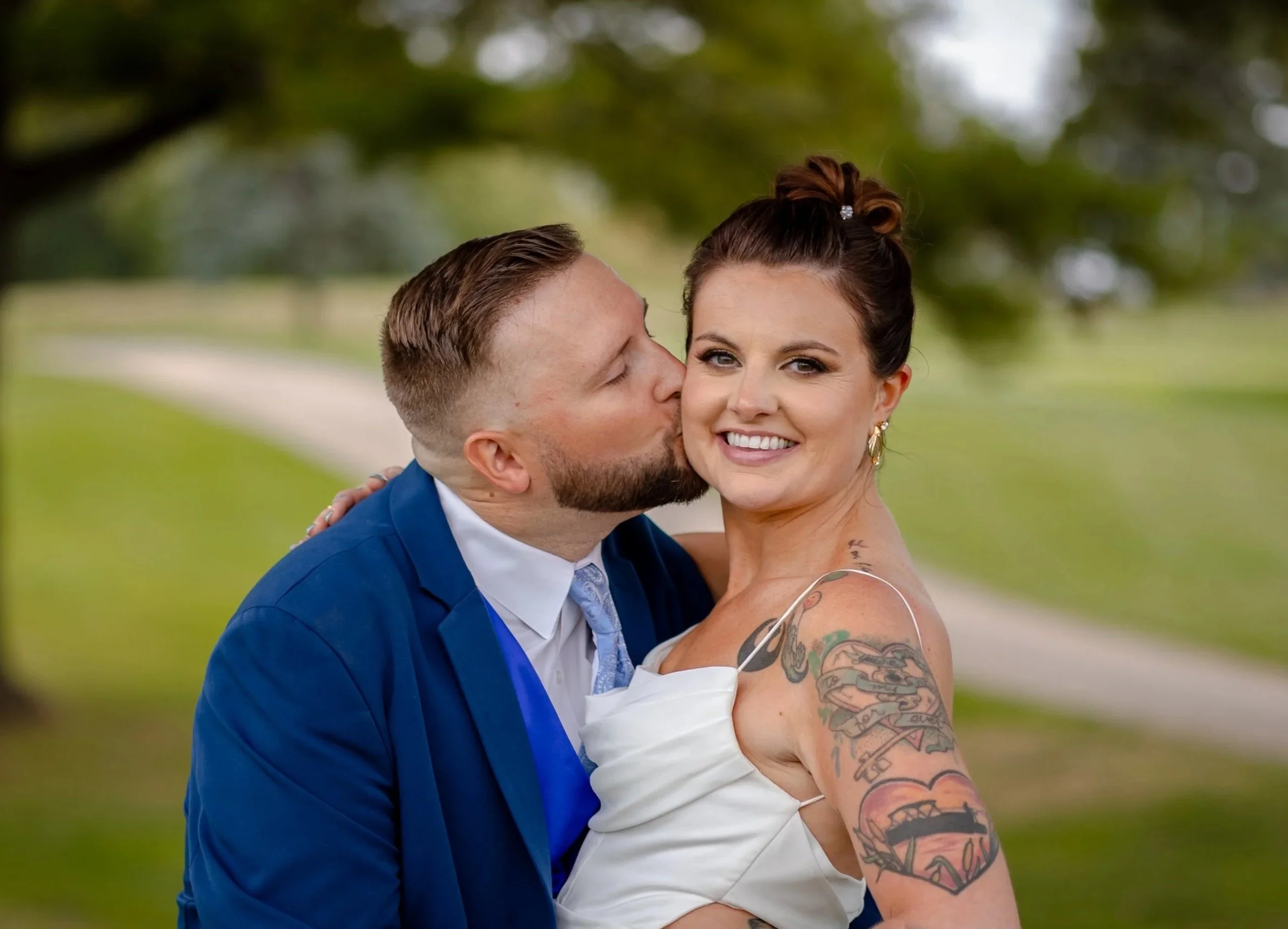 A woman and a man in wedding attire embracing outdoors, with the man kissing the woman on her cheek. The woman is smiling, has tattoos on her arm, and is wearing earrings and a wedding dress. The man is in a blue suit with a tie, and has short hair and a beard. The background is blurred with greenery.