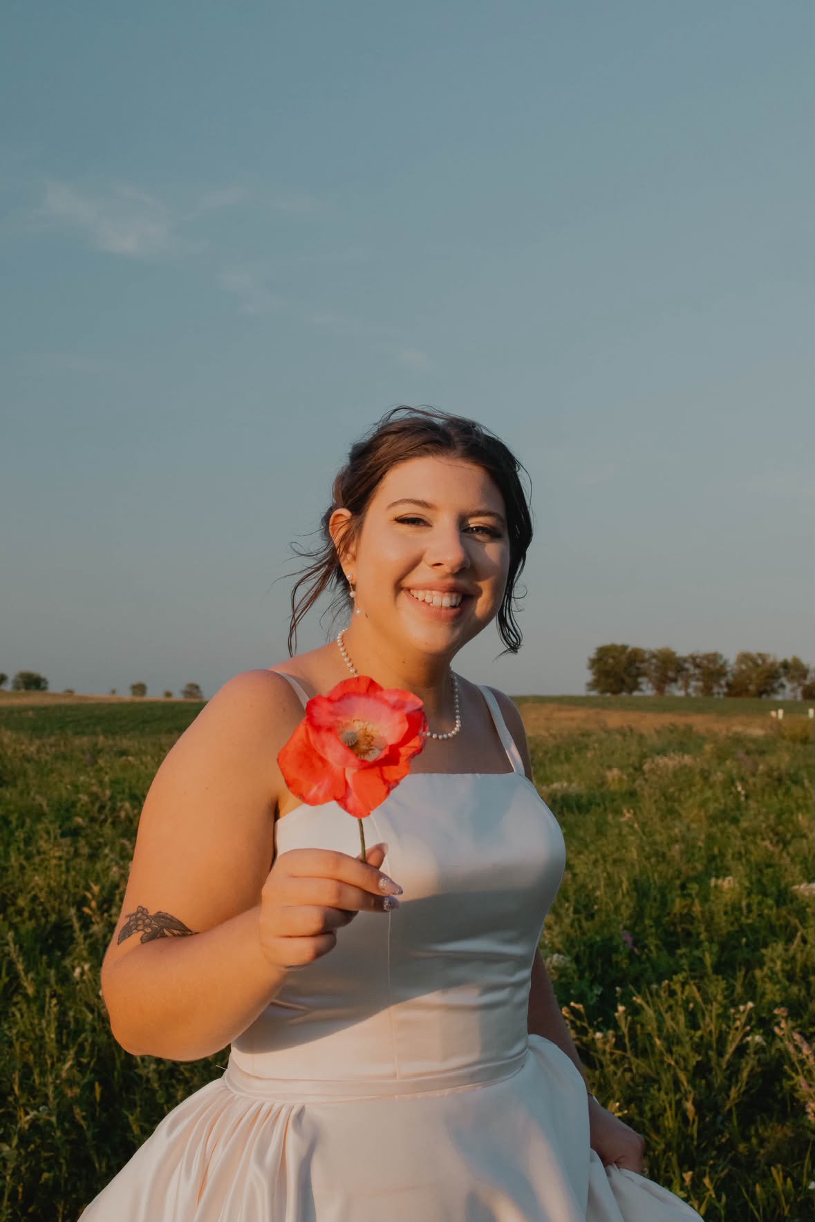 A woman in a white dress holding a red poppy flower in an open field during sunset, smiling.