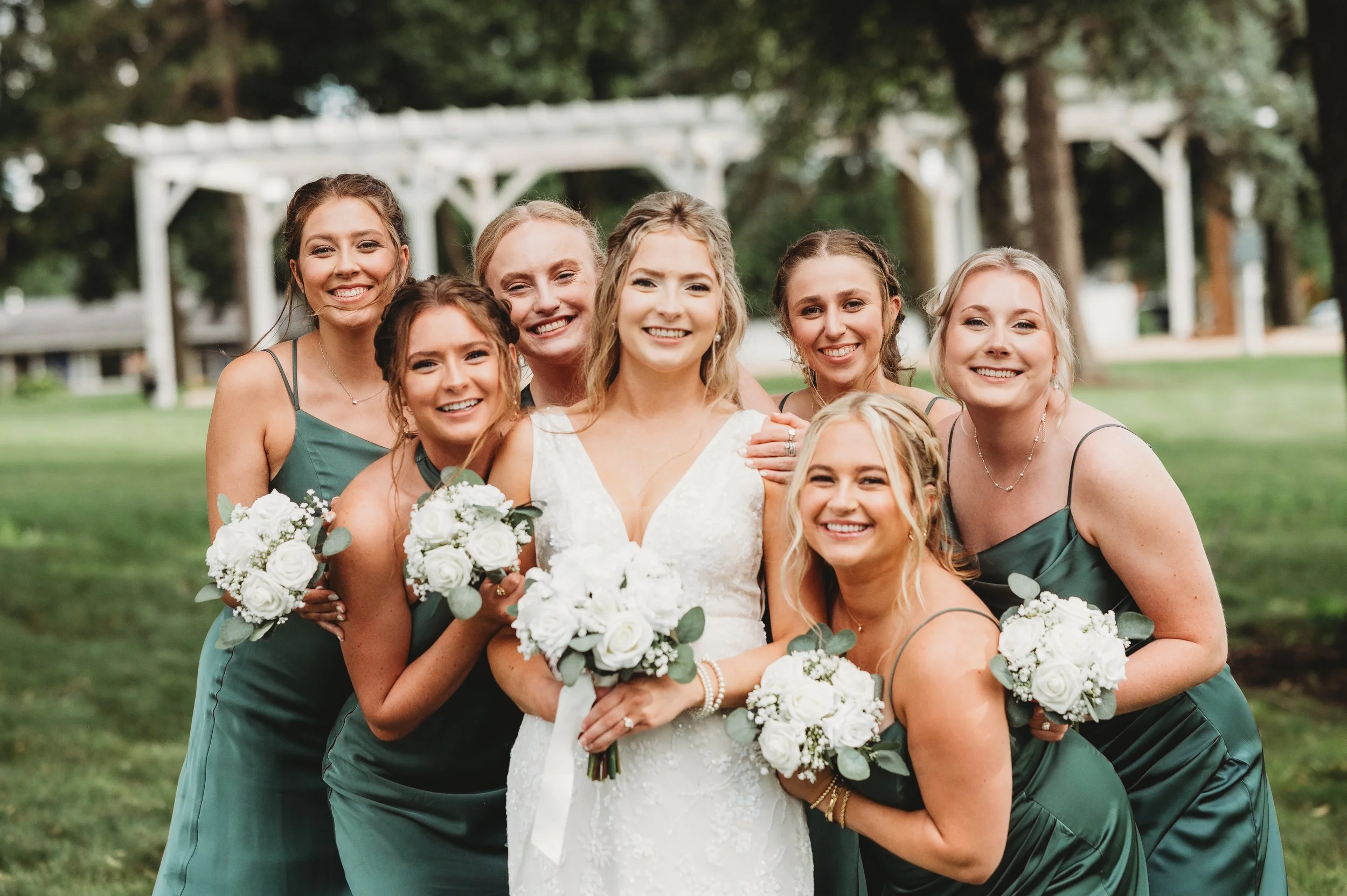 A bride in a white wedding dress holding a bouquet of white roses and surrounded by bridesmaids in dark green dresses, all smiling outdoors in a park with trees and a pavilion in the background with beautiful makeup in Madison, WI.