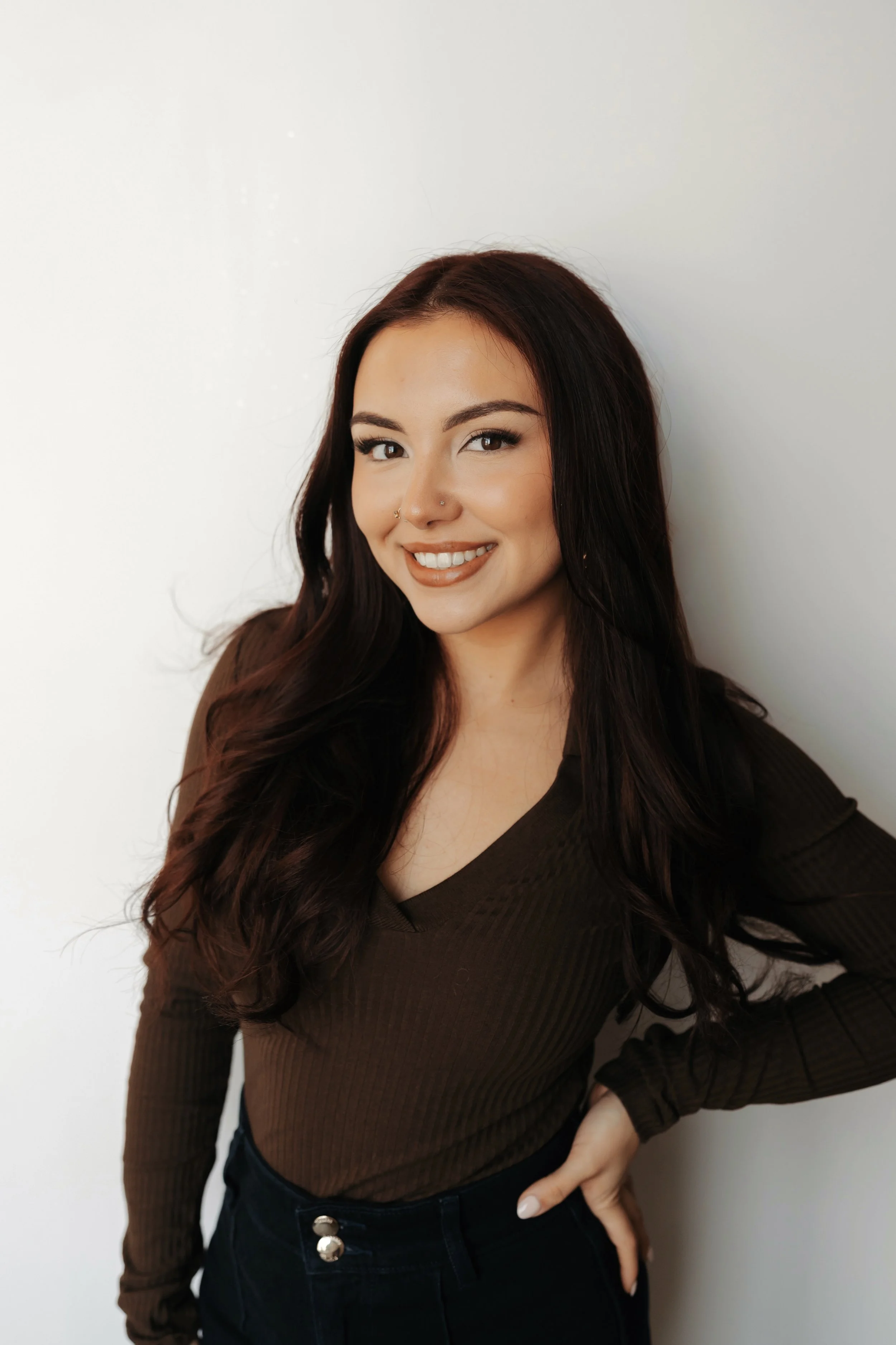 Bria with long, dark, wavy hair smiling at the camera, wearing a brown long-sleeve top and high-waisted dark jeans, standing against a plain white wall with beautiful makeup in Madison, WI