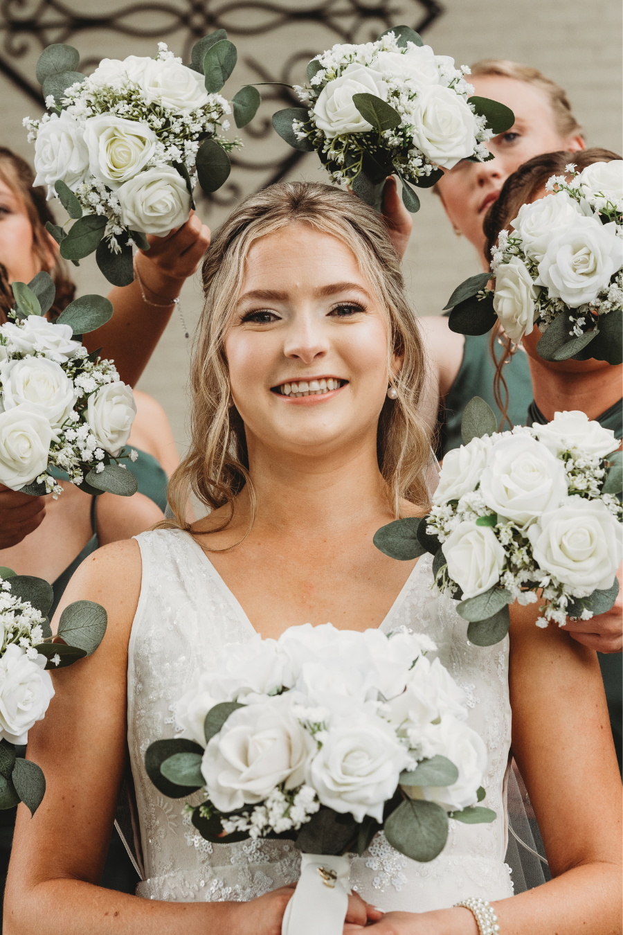 A bride smiling and holding a bouquet of white roses and greenery surrounded by bridesmaids holding similar bouquets at a wedding in Madison, WI.