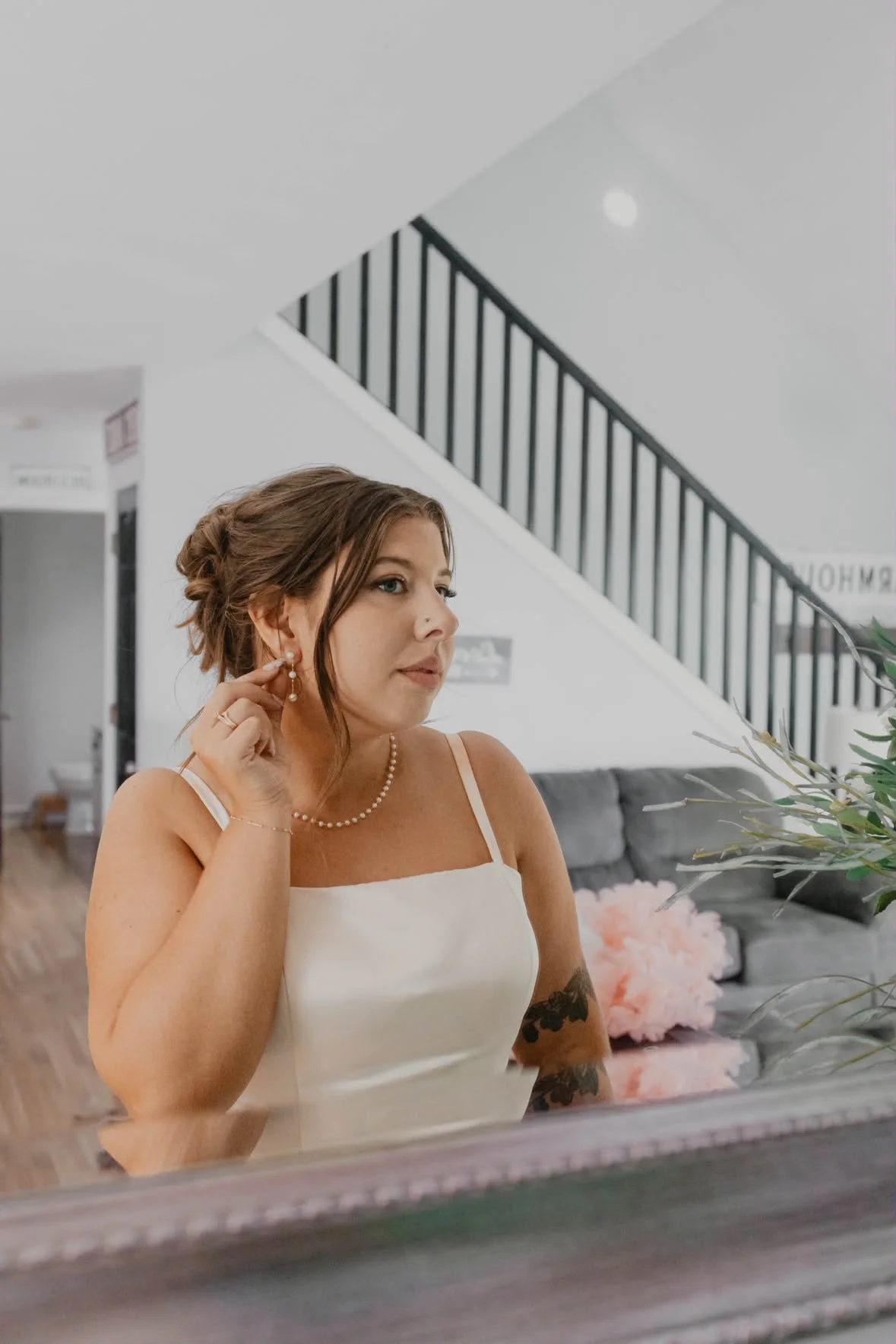 A bride with brown hair styled in an updo, wearing a wedding dress and pearl jewelry, is putting on an earring in an indoor setting with stairs and a gray couch with pink pillows in the background with beautiful makeup in Madison, WI.