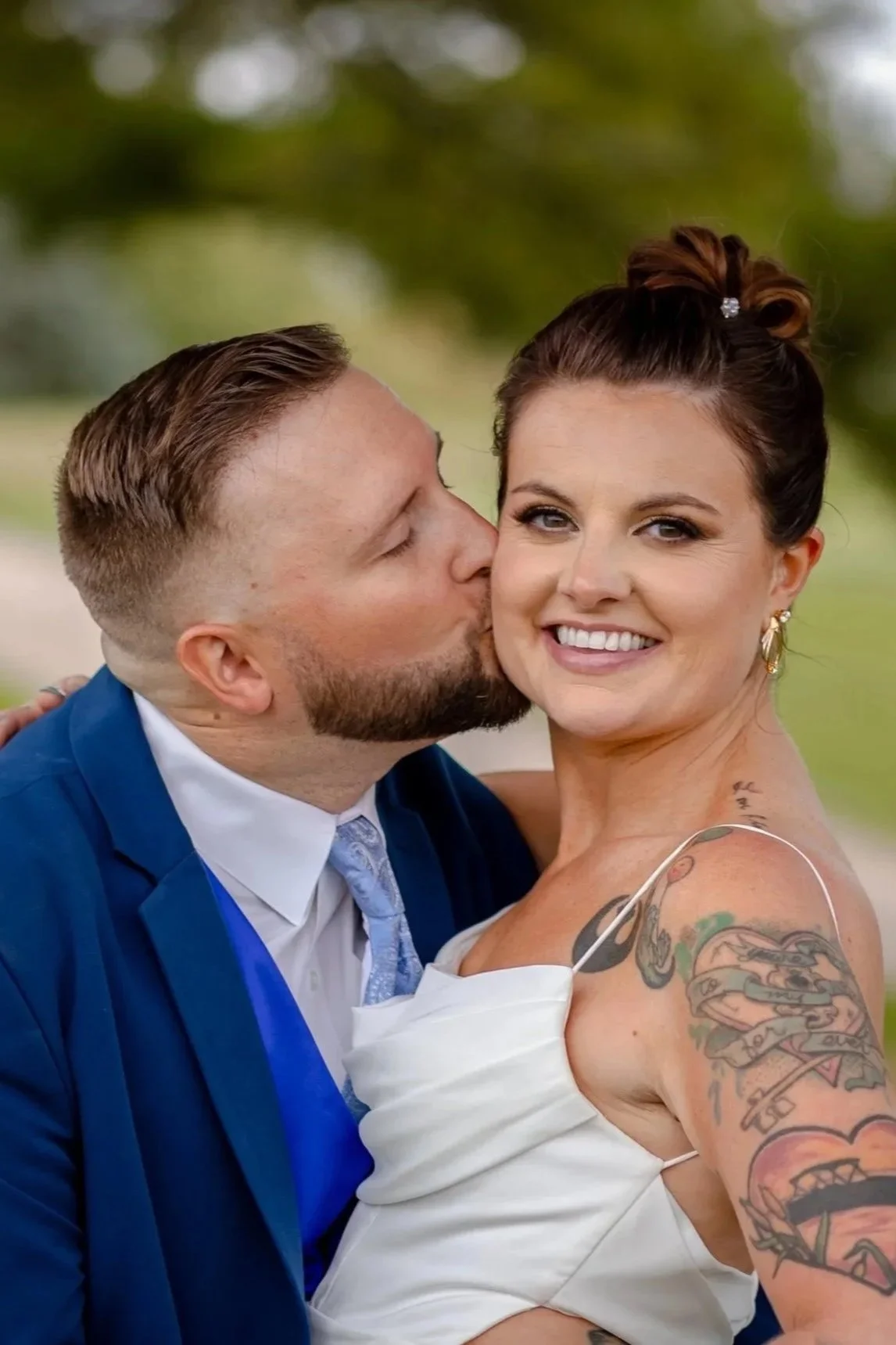 A groom in a blue suit kissing a bride in a white dress, who has beautiful makeup. The woman has tattoos on her arm and earrings, and her hair is styled up. The background is outdoor with blurred greenery in Madison, WI.