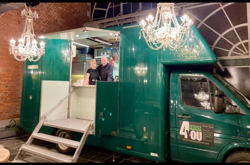 Un camion de service traiteur vert converti en stand de cuisine avec deux personnes à l'intérieur, décoré avec des lustres en cristal suspendus dans un espace intérieur.
