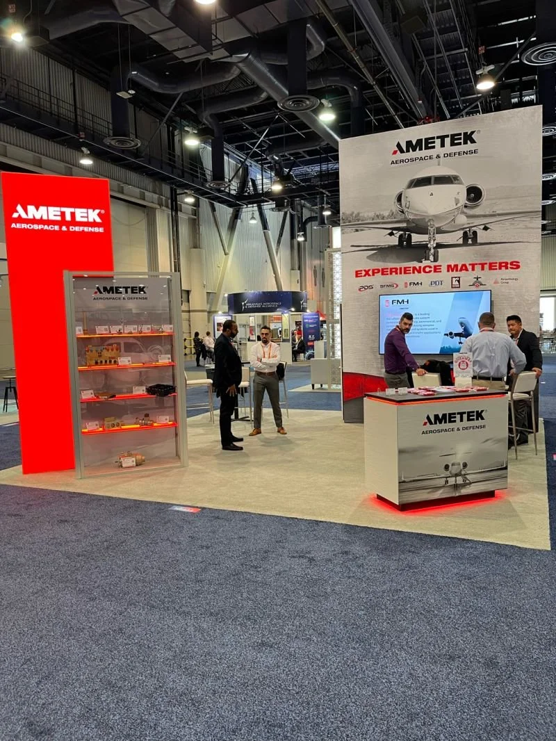 Exhibition booth for Ametek Aerospace & Defense with displays of equipment, a large sign with an airplane image, and several people talking inside the booth.
