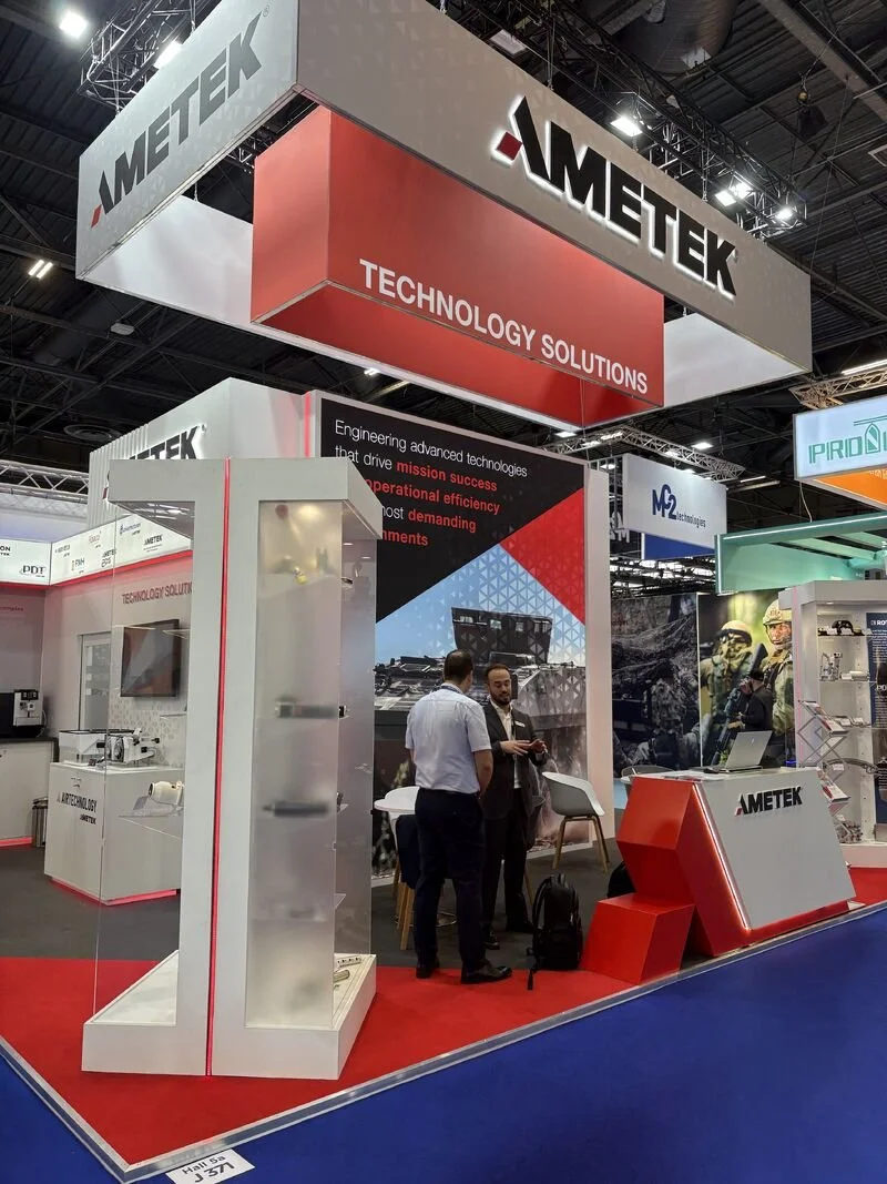 Trade show booth for AMETEK featuring red and gray color scheme, with signage displaying 'Technology Solutions' and Ametek logo, and two men conversing in front of the booth.